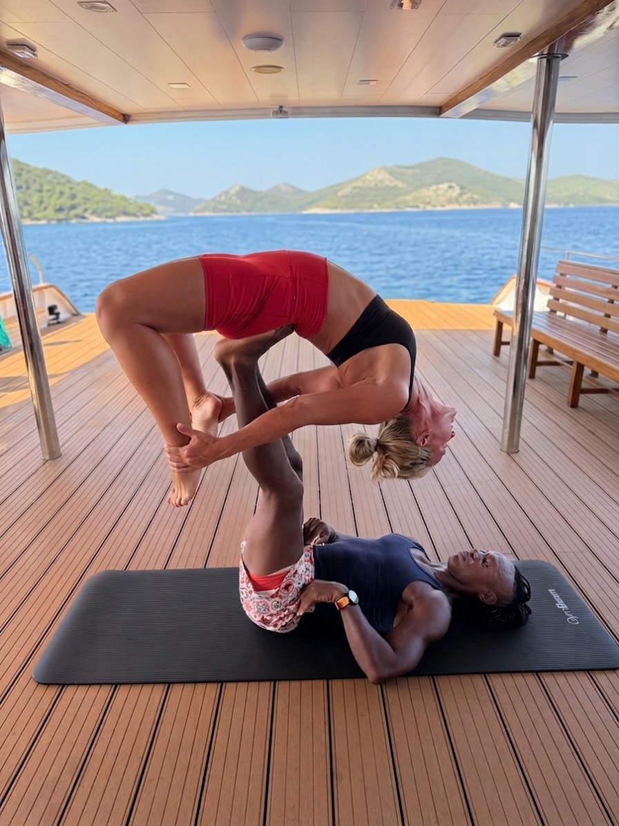 Two women practicing acro yoga on a boat deck with water and mountains in the background, one balancing on the other's hands.