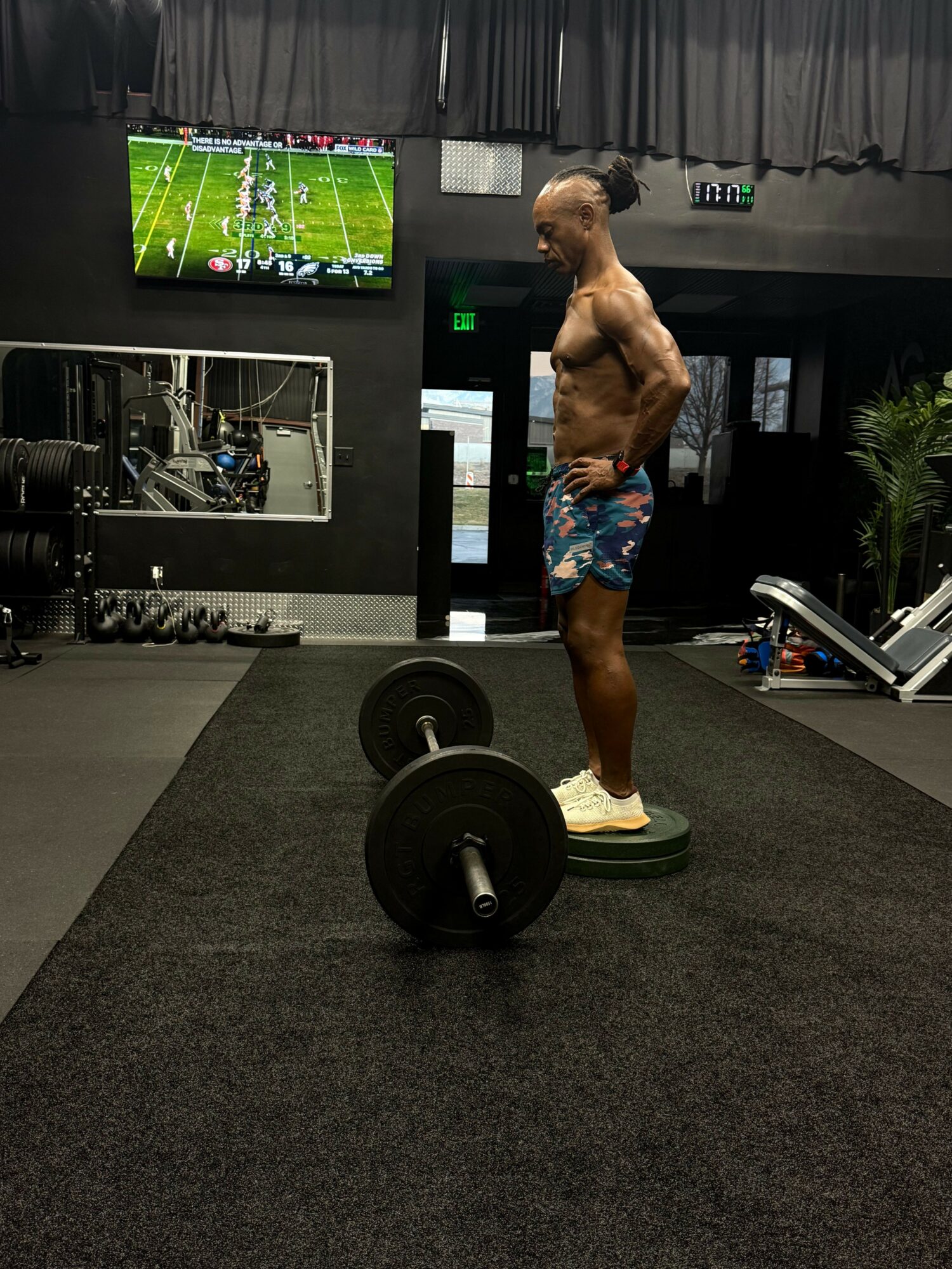 Man with a shirtless upper body and colorful shorts stands on a weight plate in a gym, with a TV and gym equipment in background.