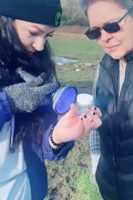 Two women outdoors, one holding a small container, both looking at it, with grass and trees in the background.