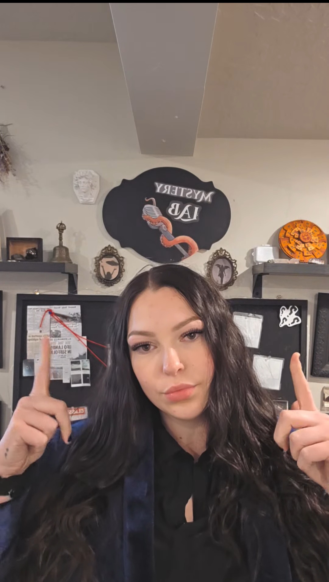 Young woman with long dark hair pointing upward, indoor background with decorations and a Halloween pumpkin.