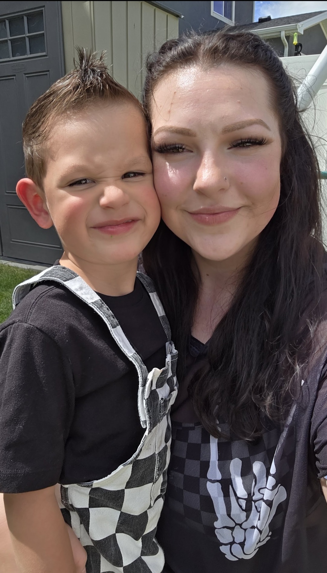 A woman and a young boy smiling outdoors, close together, with a house in the background.