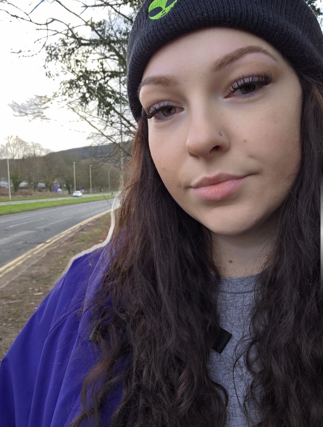 Close-up of a young woman with long dark hair wearing a black beanie and purple jacket outdoors.