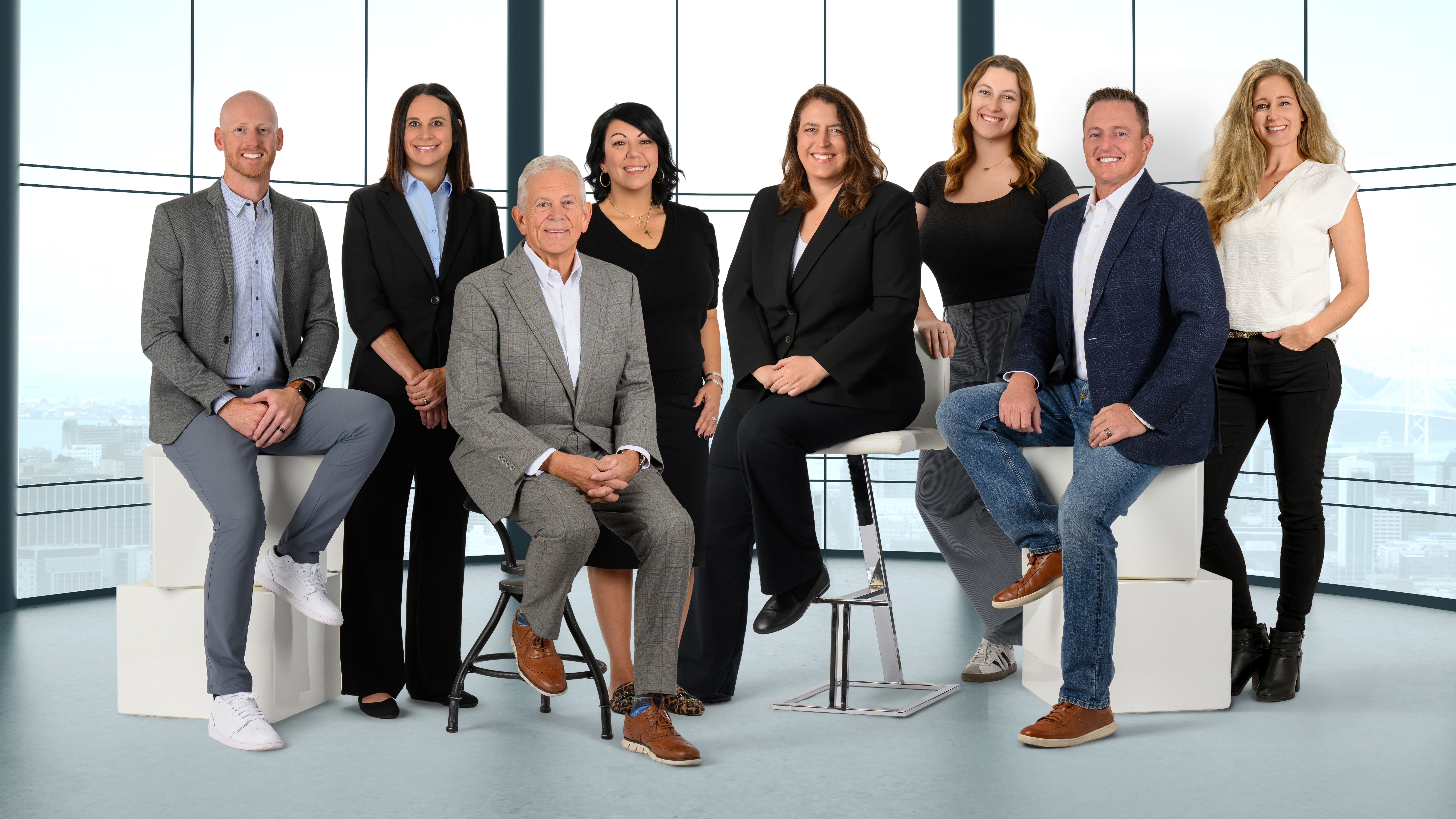 Group of nine diverse professionals in a modern office, some seated and some standing, with large windows in the background.