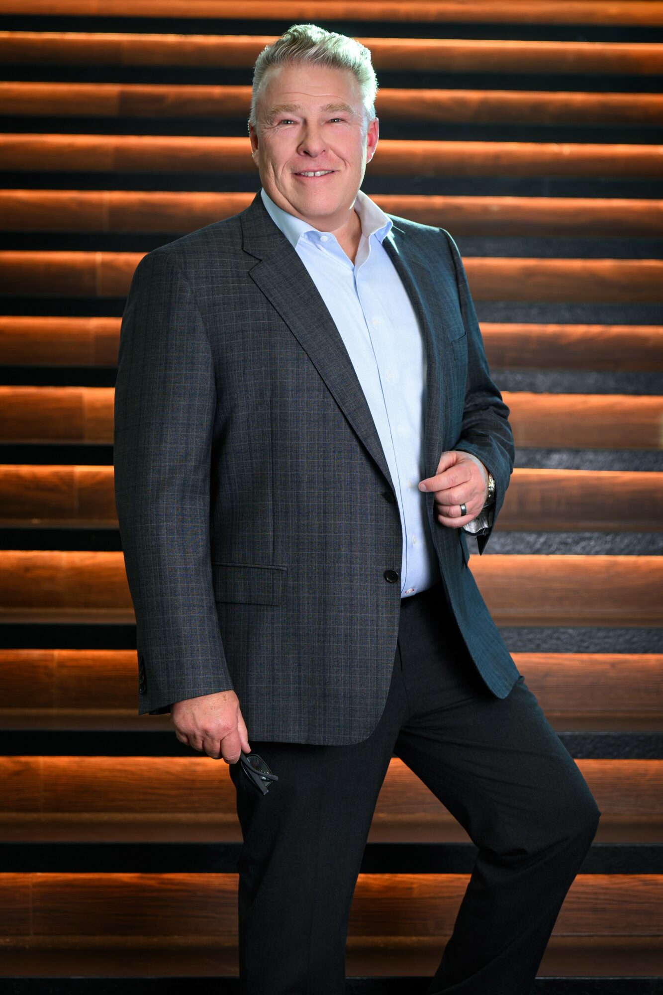 Man in a suit standing on wooden stairs, smiling, holding a phone, with a dark background.