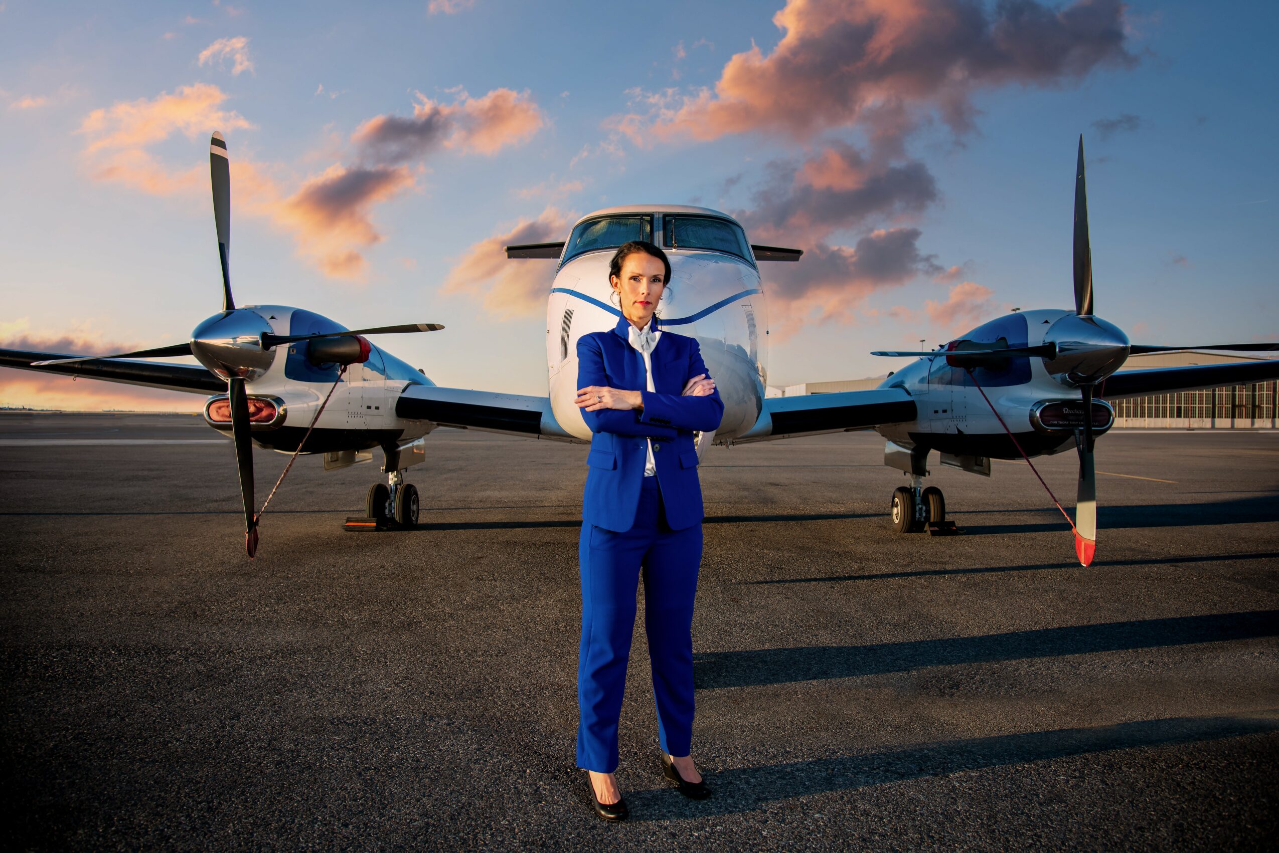 Woman in blue jumpsuit standing in front of a small airplane on runway at sunset.
