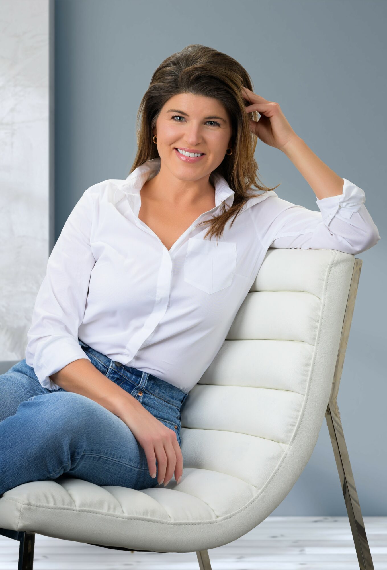 Woman with brown hair smiling, sitting on a white chair, wearing a white shirt and blue jeans, in a bright room.