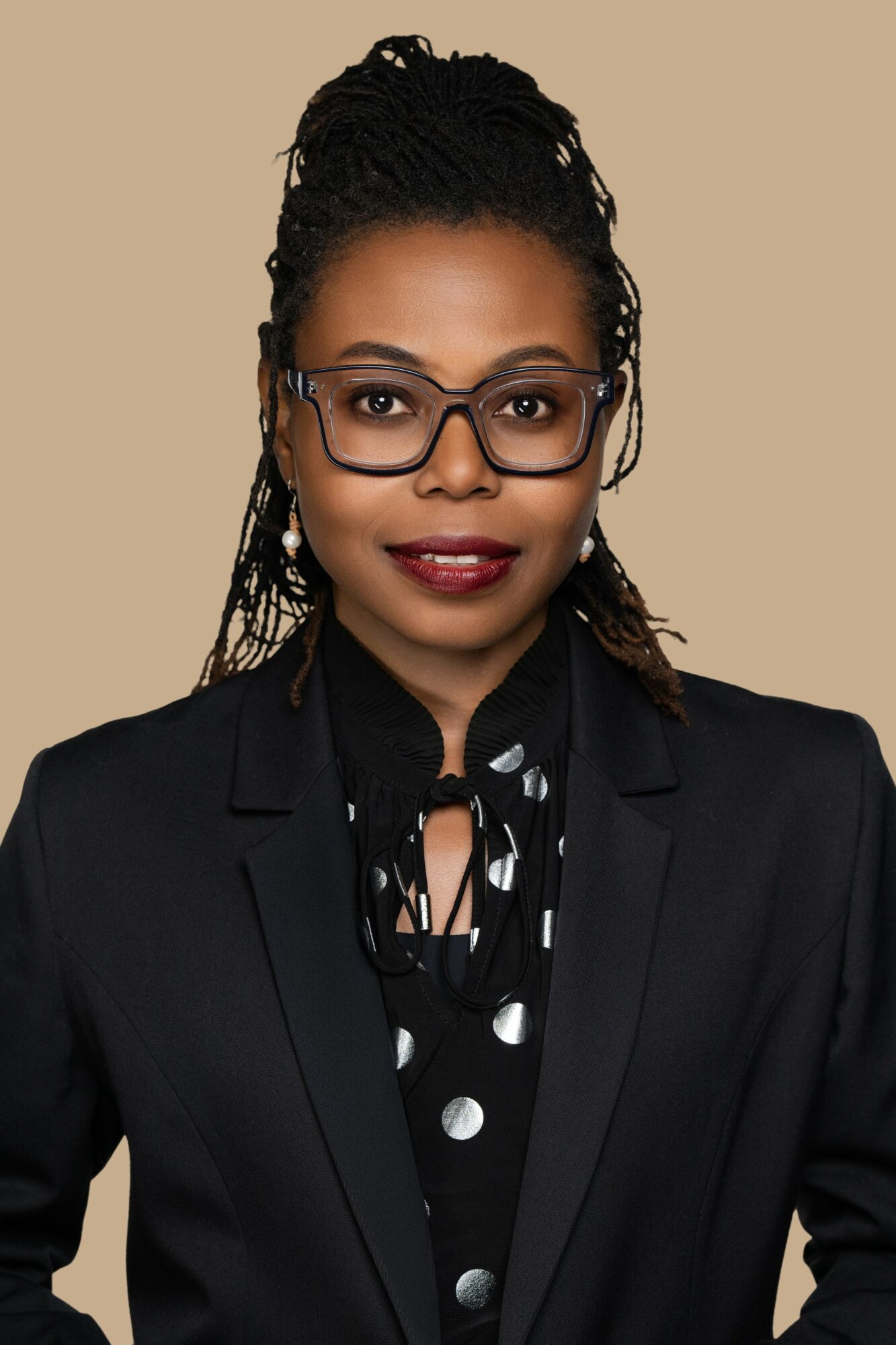 Woman with glasses and braided hair wearing a black blazer and polka dot blouse against beige background.