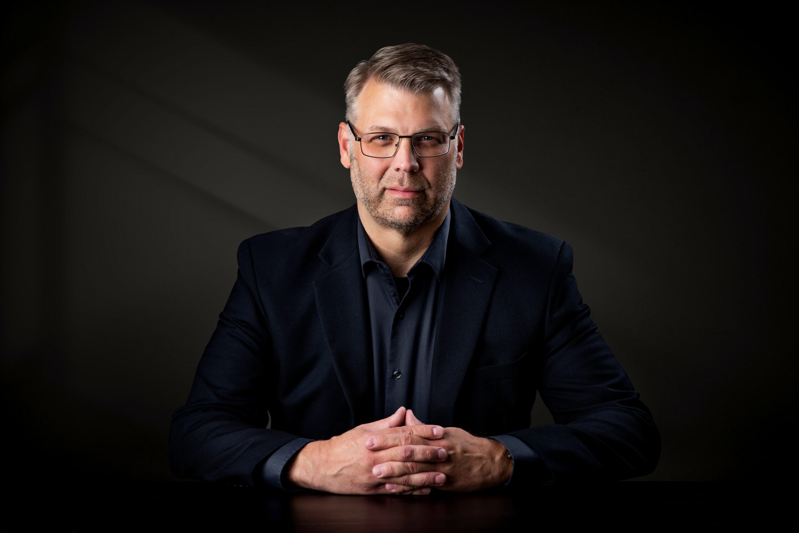 Man with glasses and beard in dark suit and shirt, sitting with hands clasped, against dark background.