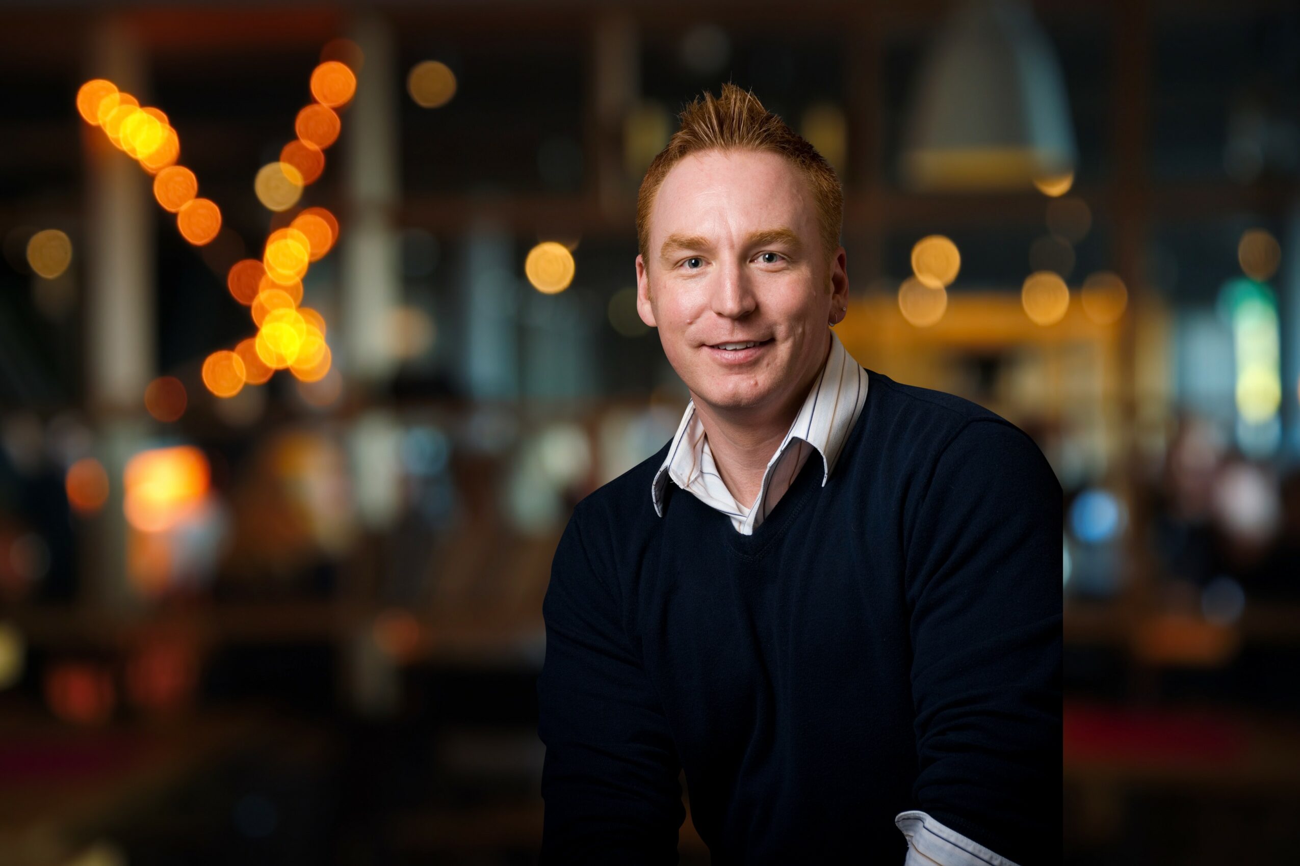 Man with short hair and a dark sweater sitting in a dimly lit restaurant or bar, smiling at the camera.