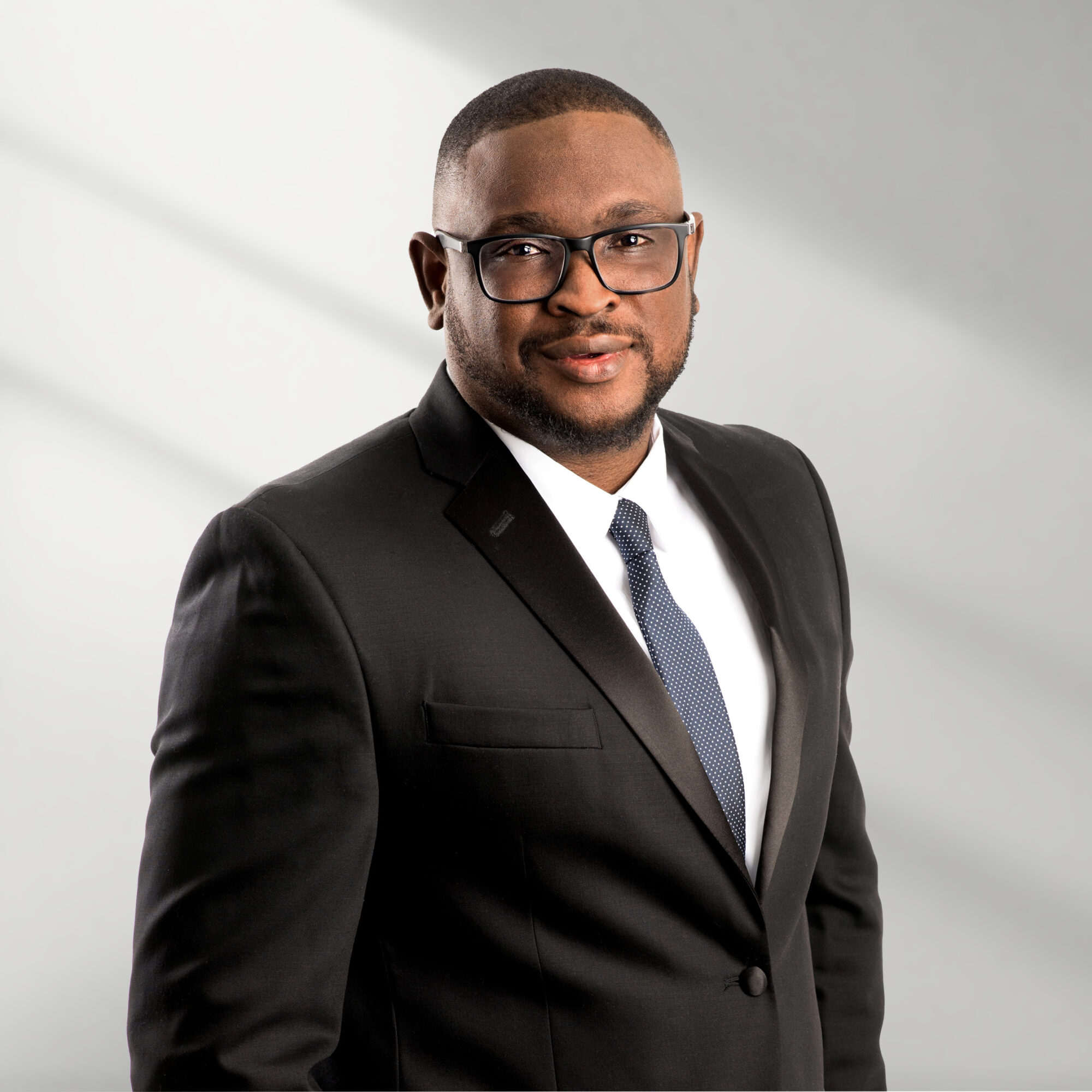 Man in black suit, white shirt, and tie wearing glasses, standing against a light background.