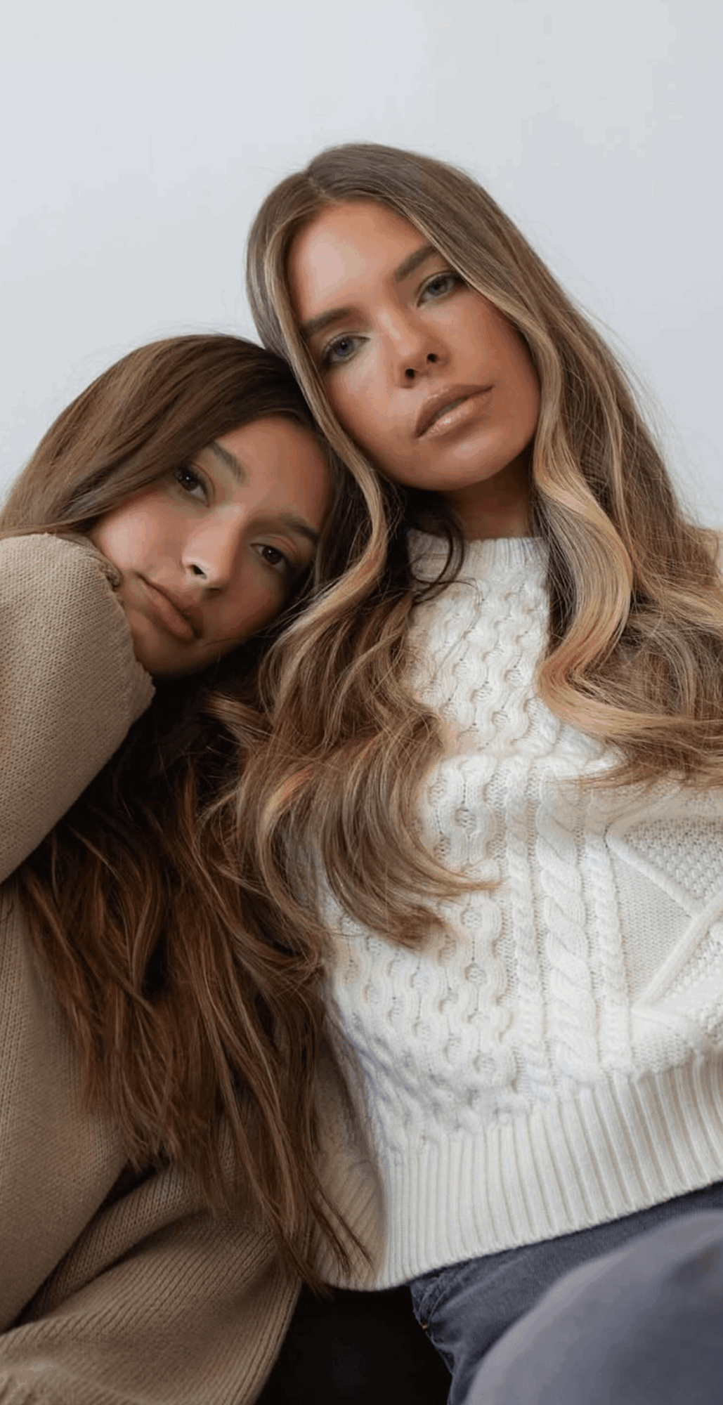 Two young women with long hair, one resting her head on the other's shoulder, both looking at the camera.