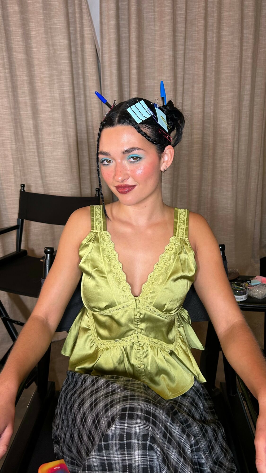 Young woman with dark hair in buns, wearing a lime green top and plaid skirt, sitting indoors with curtains behind her.