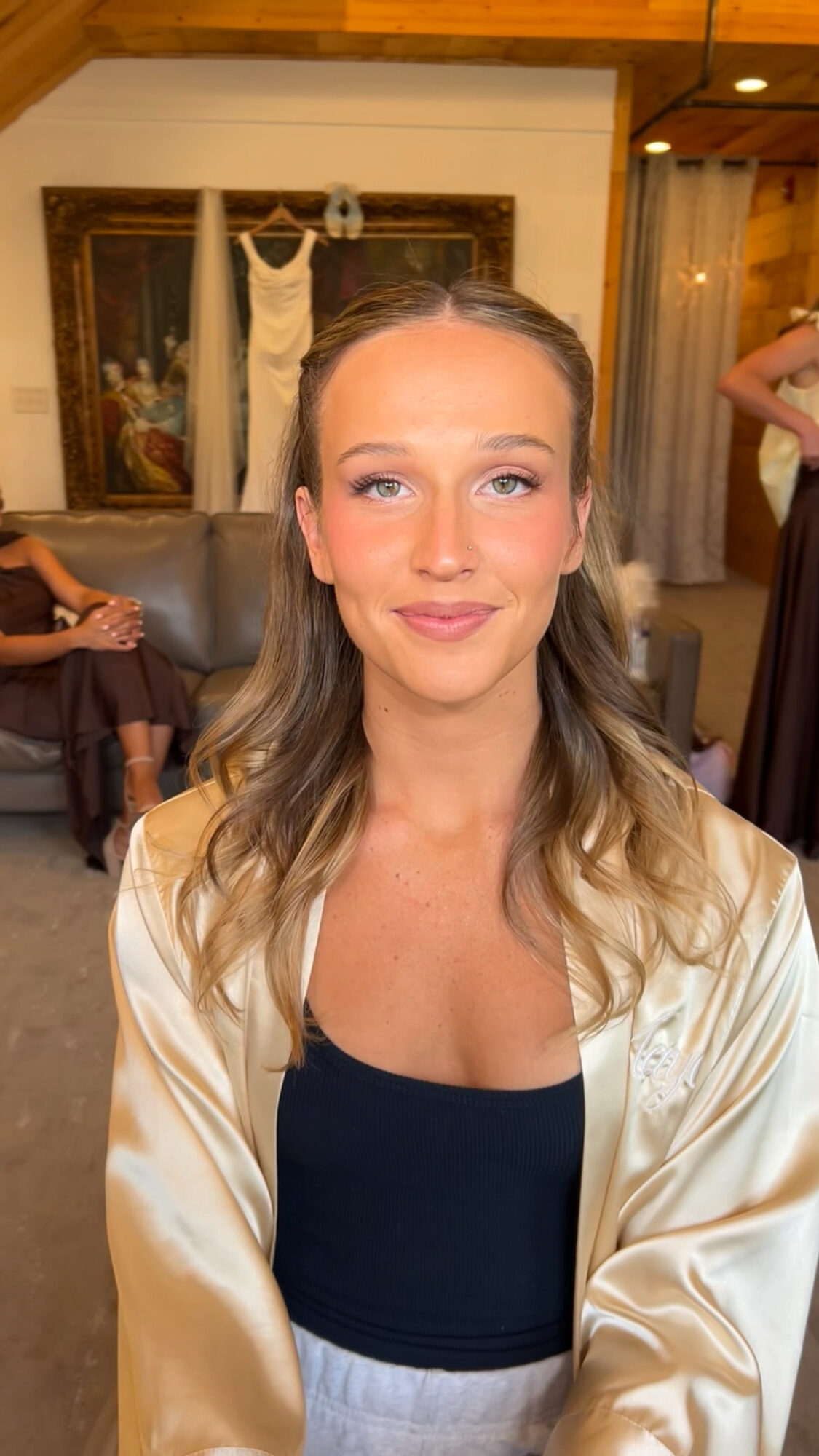 Young woman with wavy hair and light makeup, wearing a black top and cream-colored satin robe, smiling indoors.