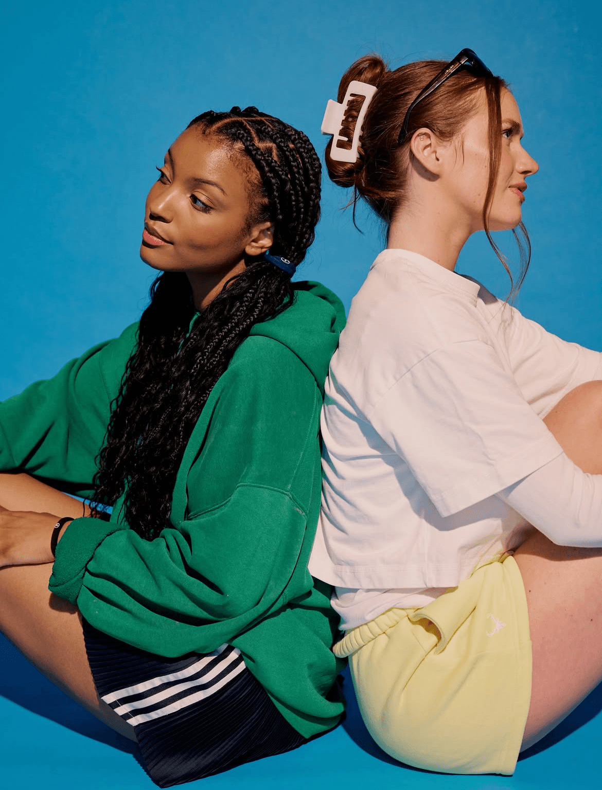 Two women sit back-to-back against a blue background, one in a green hoodie and the other in a white top and yellow shorts.