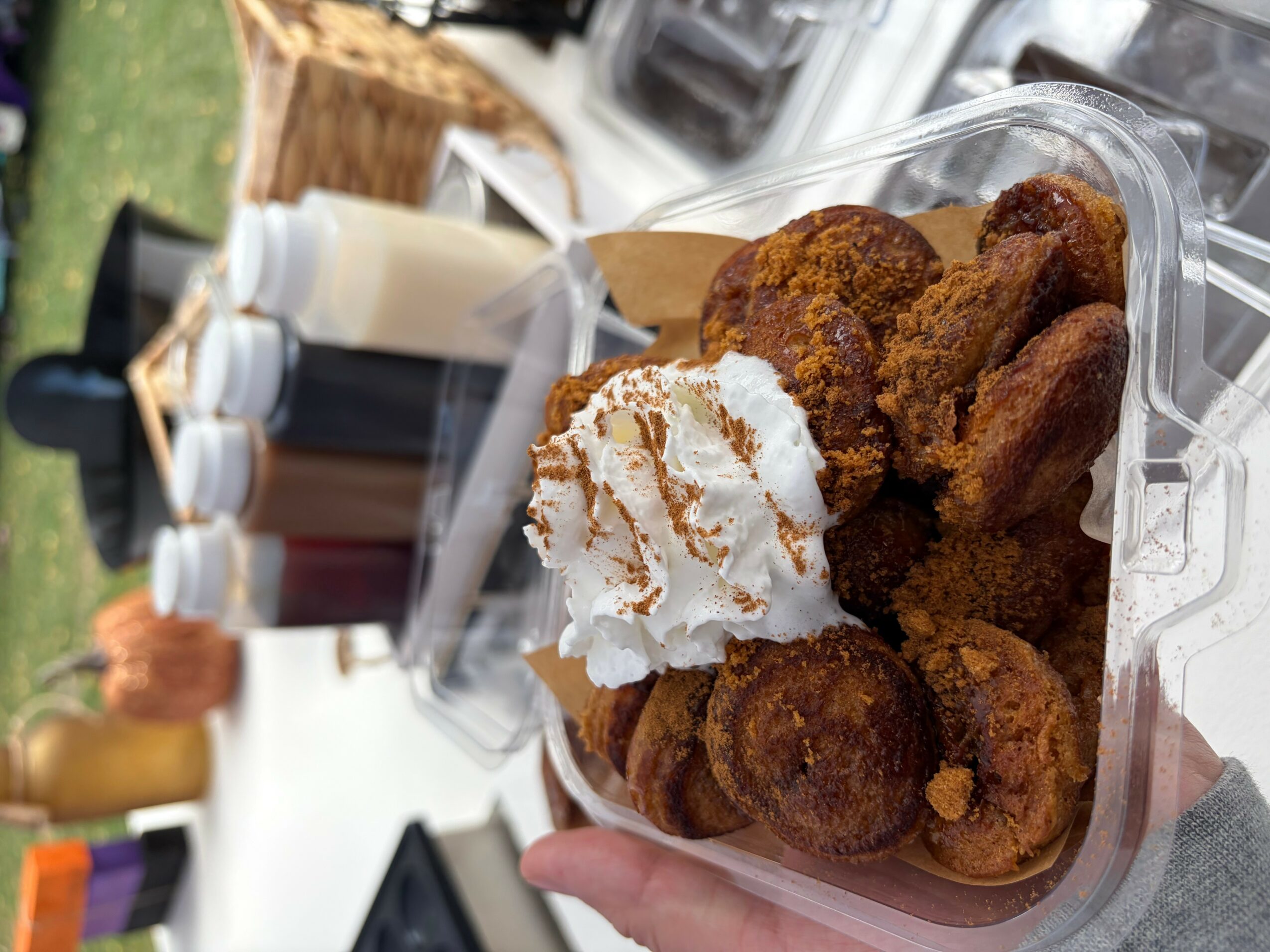 Hand holding a container of fried food with whipped cream, with bottles and outdoor setting in background.