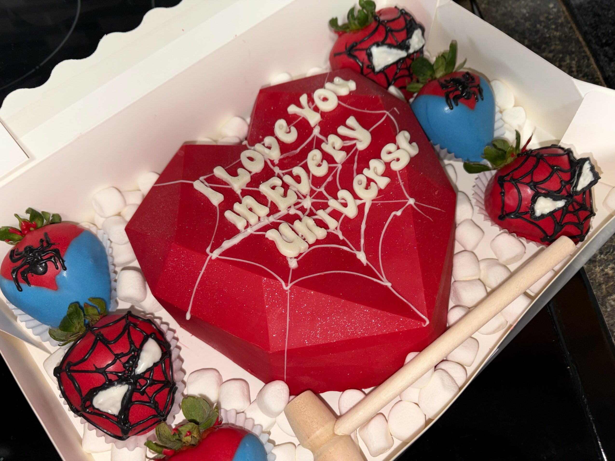 Heart-shaped cake with white writing, surrounded by decorated strawberries in a box, with white padding, on a dark surface.