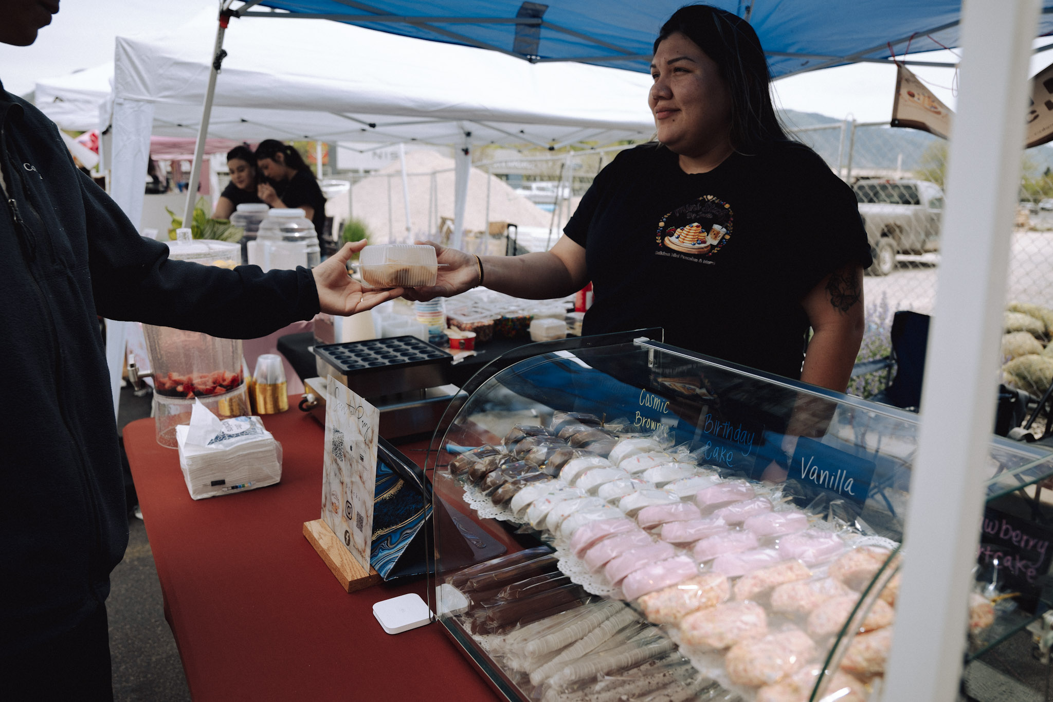 A woman at a market stall hands a customer a pastry or snack, with a display of baked goods in front. The stall has a blue canopy.