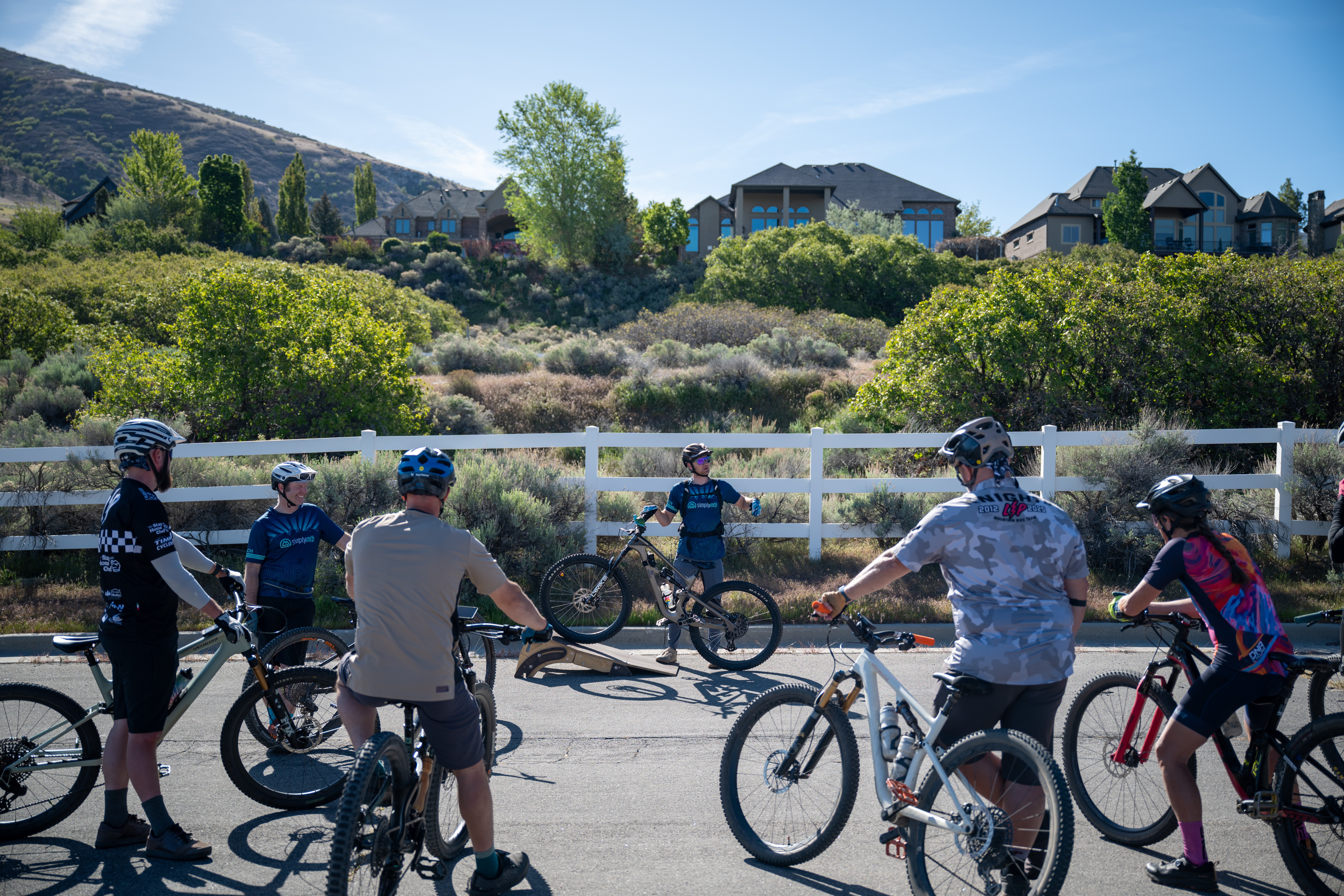 Group of children and adults with bicycles standing outdoors near a white fence, with houses and greenery in the background.