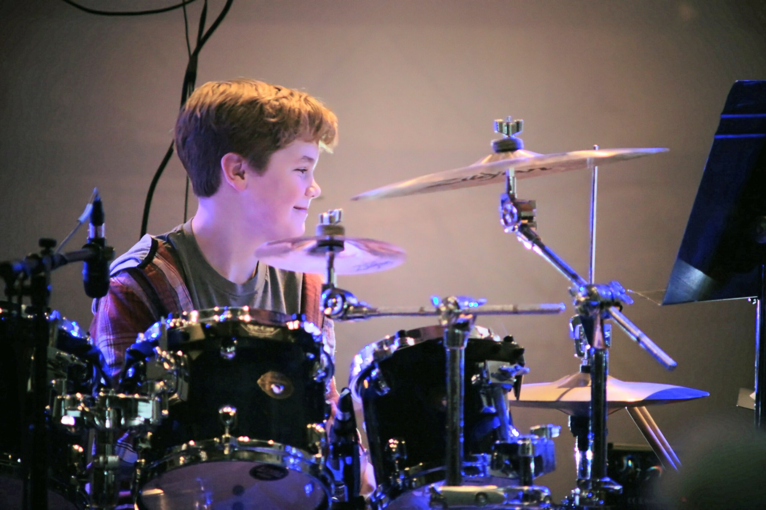 Young boy playing drums on stage, smiling, with drum set and cymbals visible, and a monitor screen nearby.