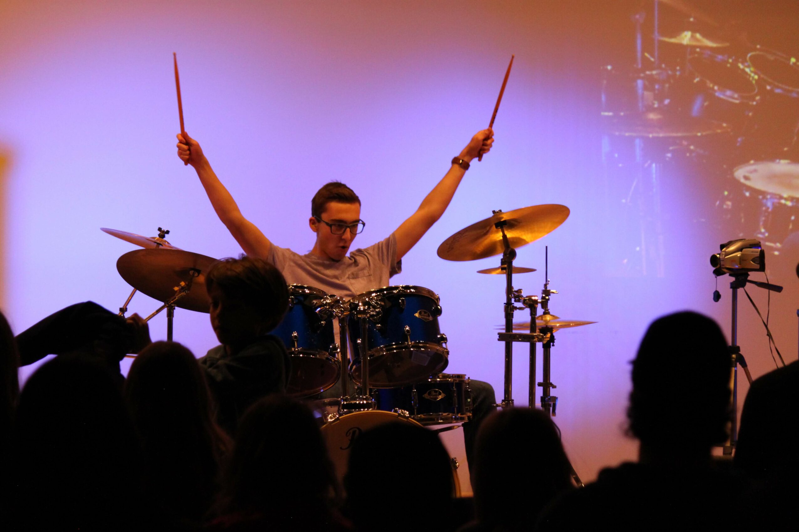 Musician playing drums on stage with arms raised, audience silhouettes in foreground, purple background, stage equipment visible.