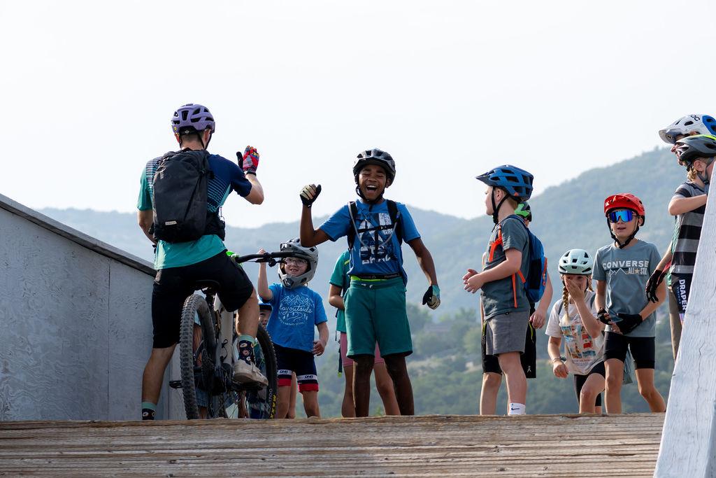 Group of children and adults with bicycles outdoors, some wearing helmets, standing on a wooden platform near a railing, with hills in the background.