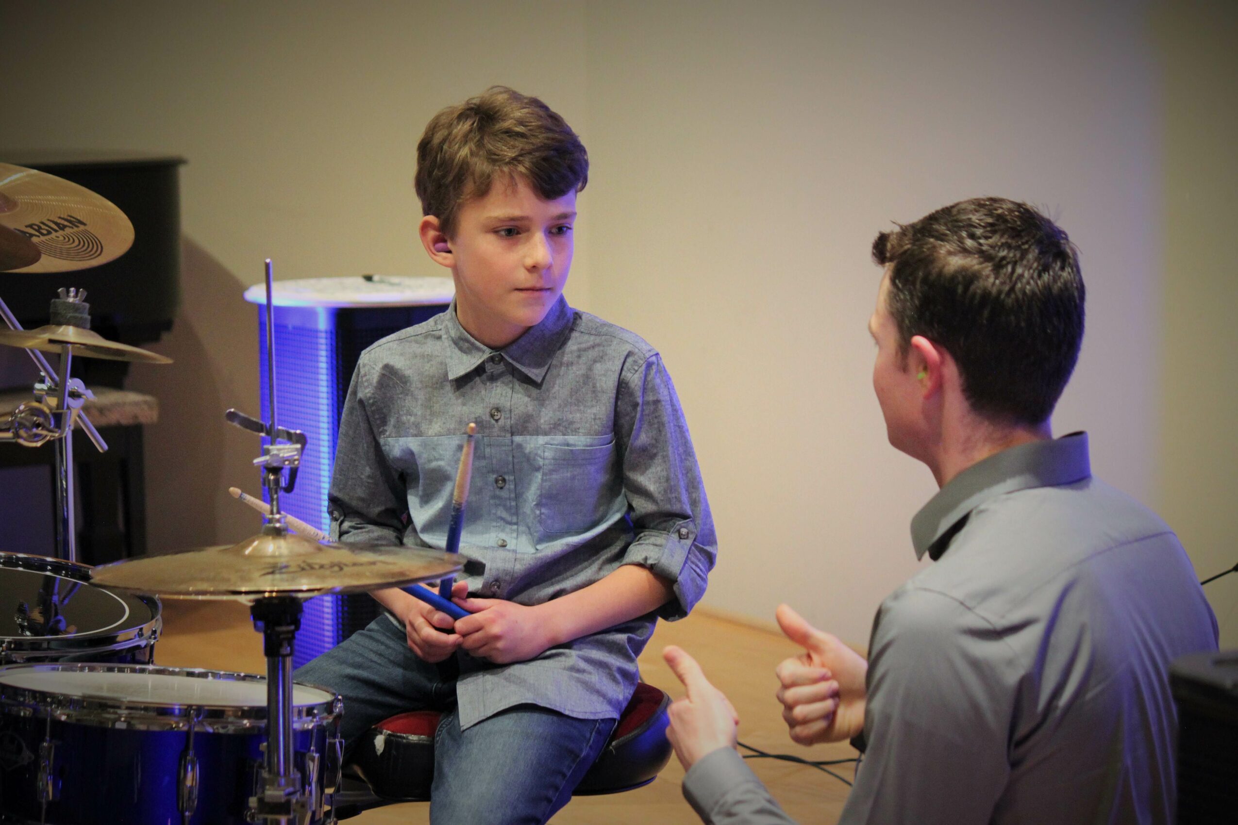 Two boys sitting and talking, one holding drumsticks, in a room with musical instruments, neutral background.
