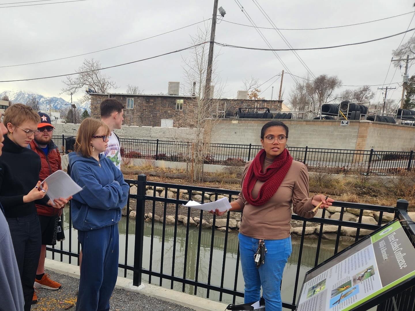 Group of people standing outdoors near a black fence, with a woman speaking and holding papers, others listening attentively.