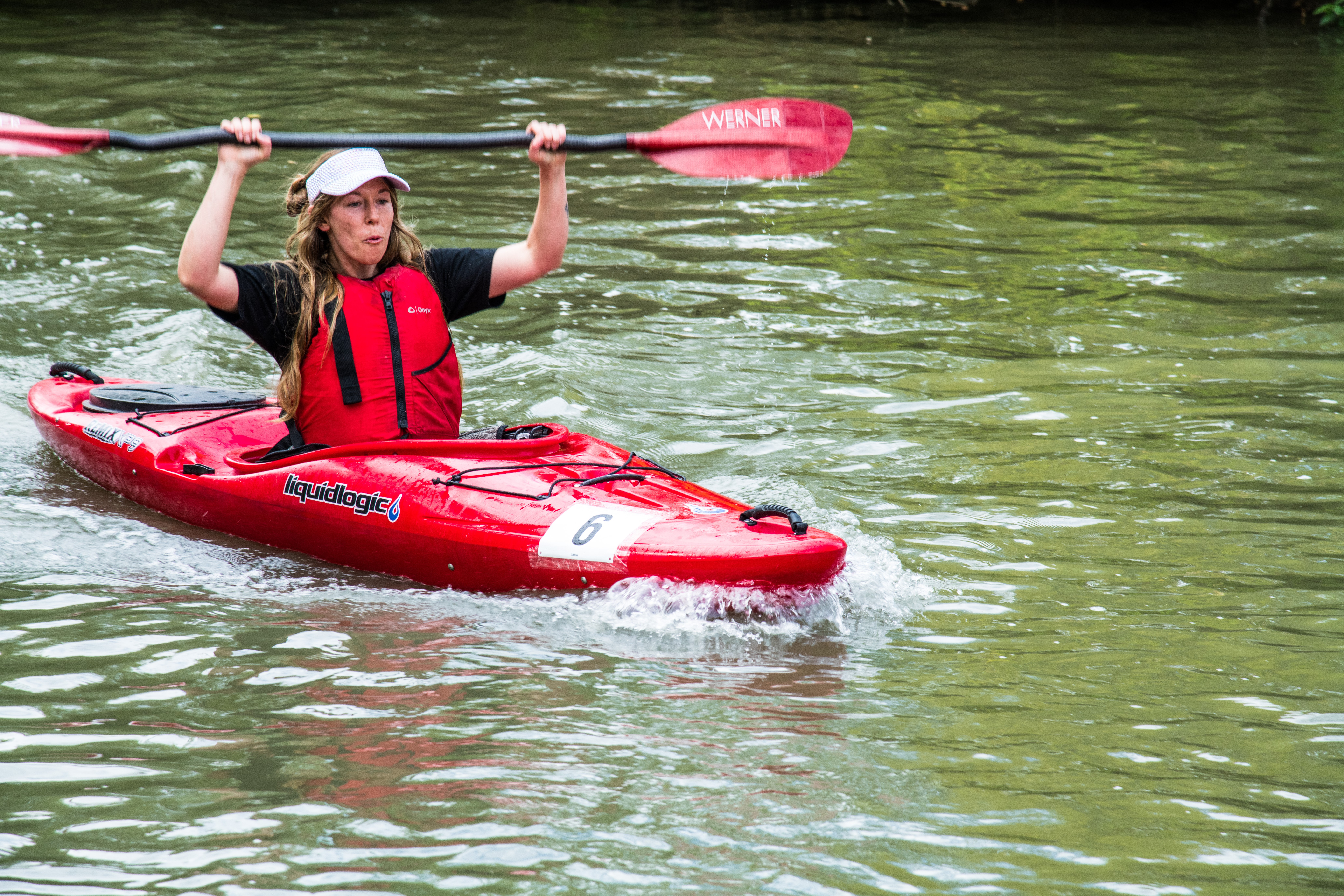 Person kayaking on water, holding paddle above head, wearing life jacket and hat, surrounded by green water.