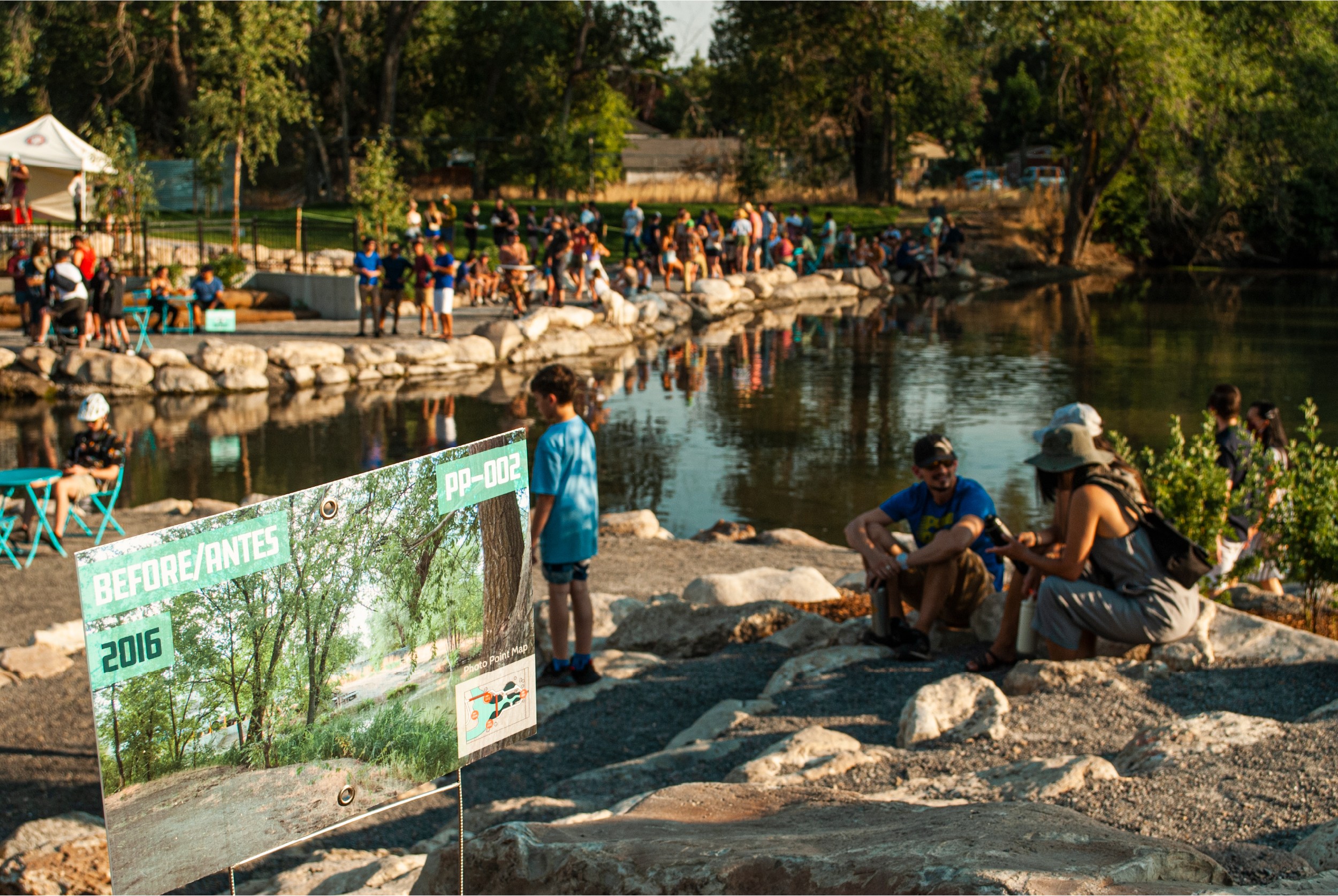 People gather along a riverbank with trees and a crowd in the background, some sitting on rocks and others walking.