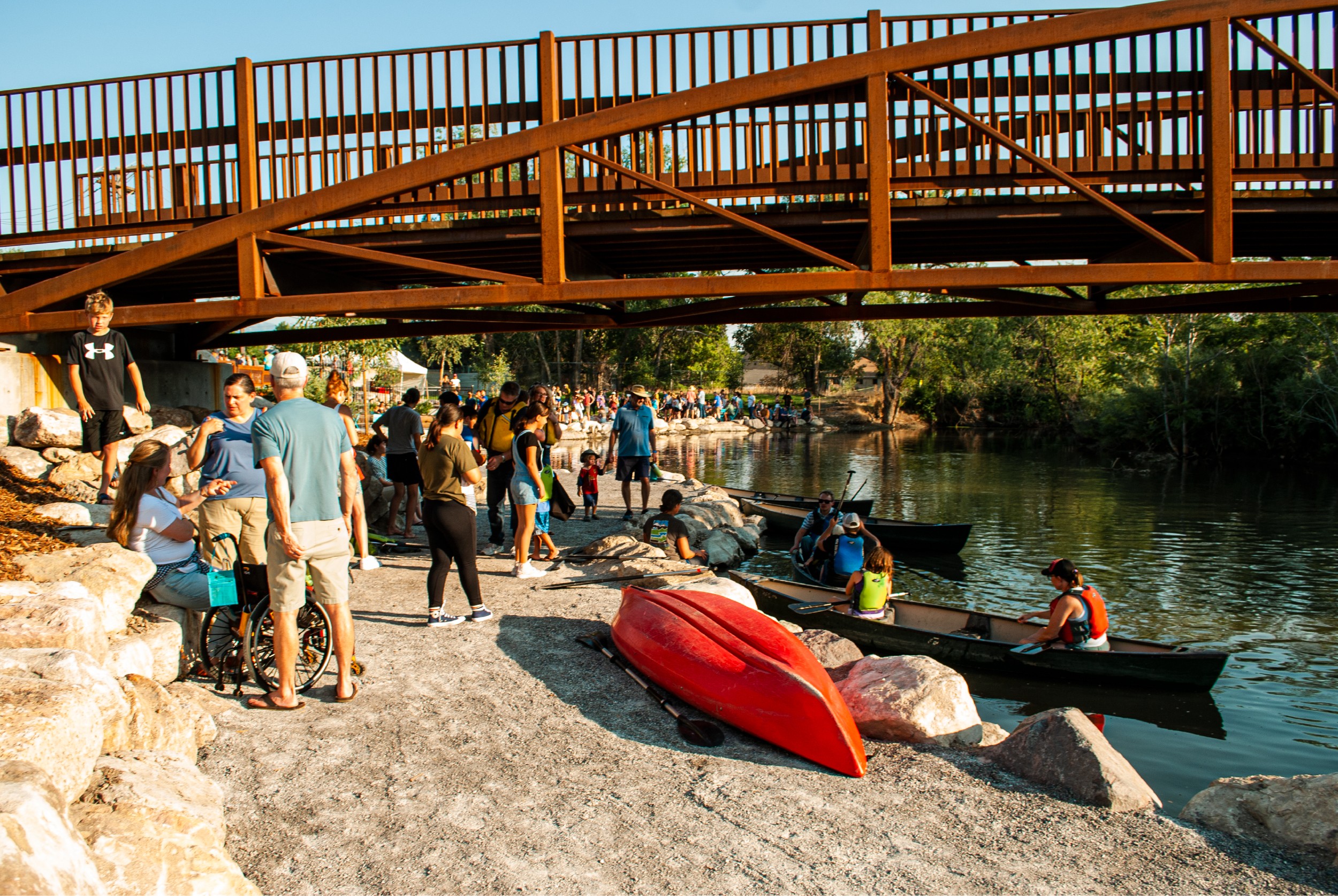 People gather near a river with boats, a red kayak, and a wooden bridge overhead on a sunny day.