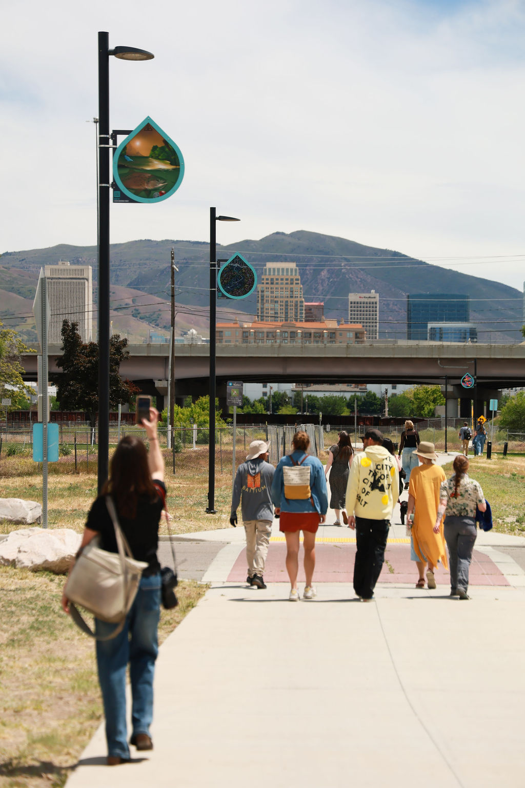 People walking on a sidewalk with city buildings and mountains in the background, under cloudy sky.