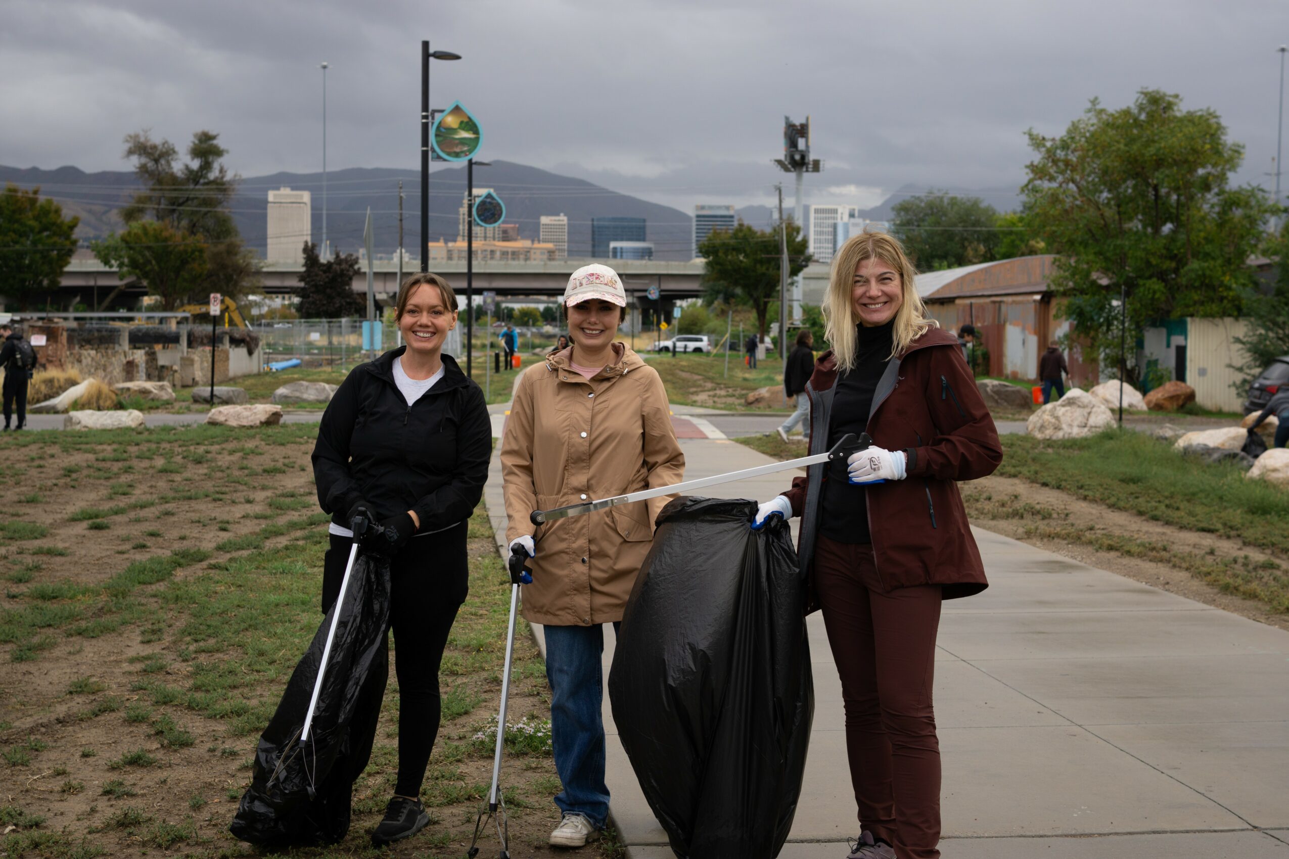 Three women standing outdoors on a sidewalk, smiling, with a cityscape and cloudy sky in the background.