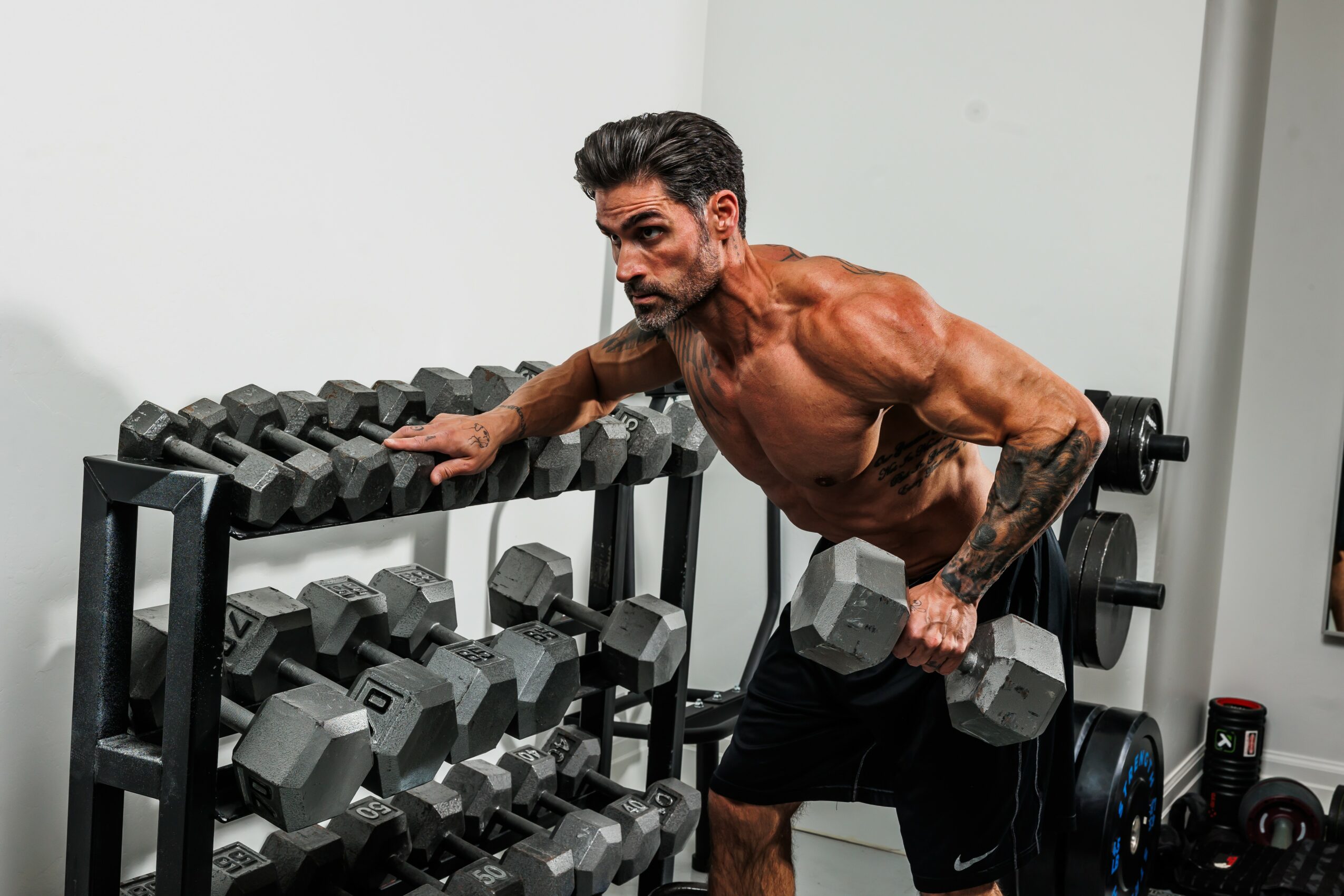 A muscular man with gray hair and beard lifts a dumbbell in a gym, surrounded by dumbbells and workout equipment.