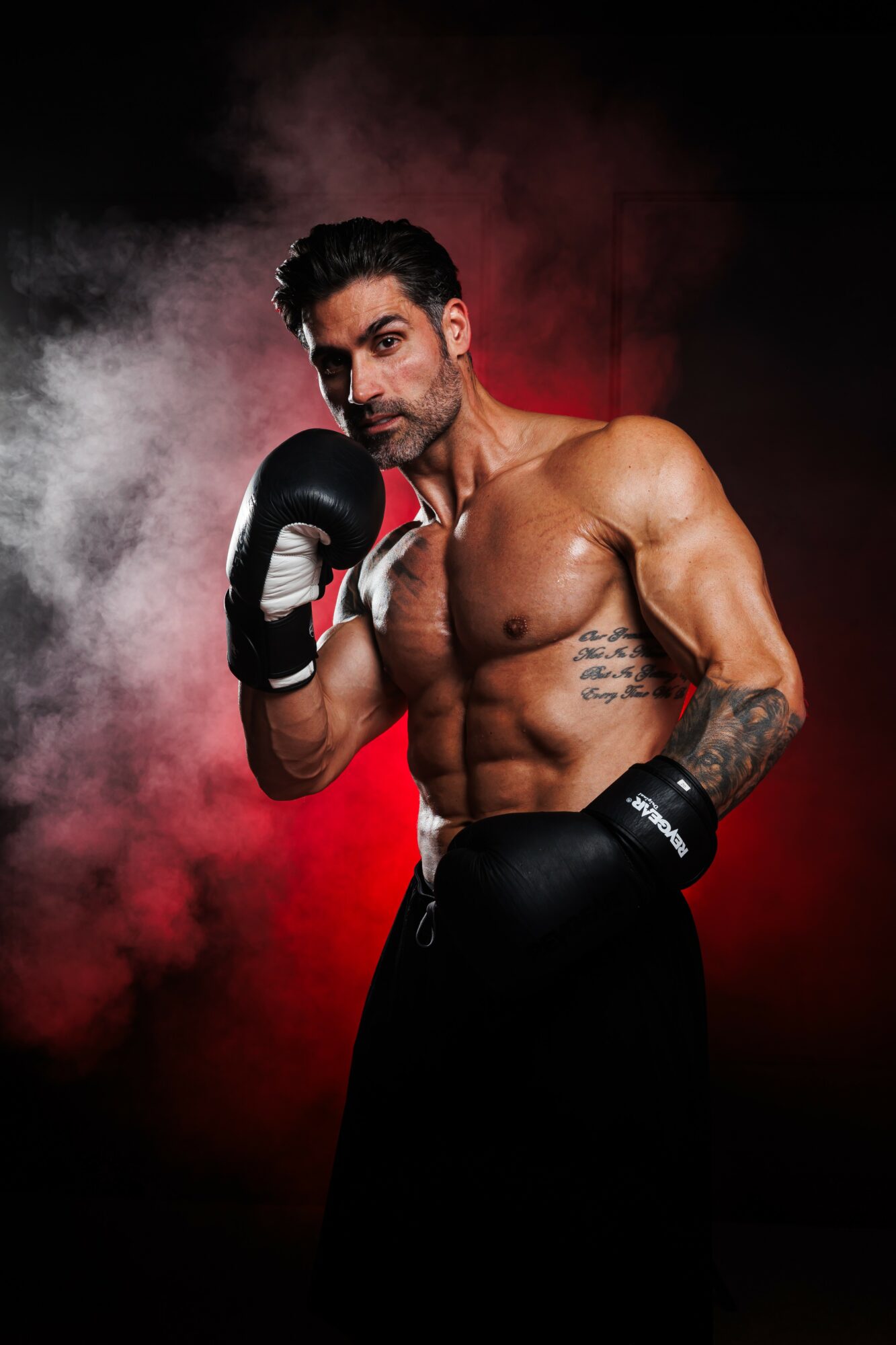A shirtless male boxer with gloves, standing in a boxing stance against a smoky red and black background.