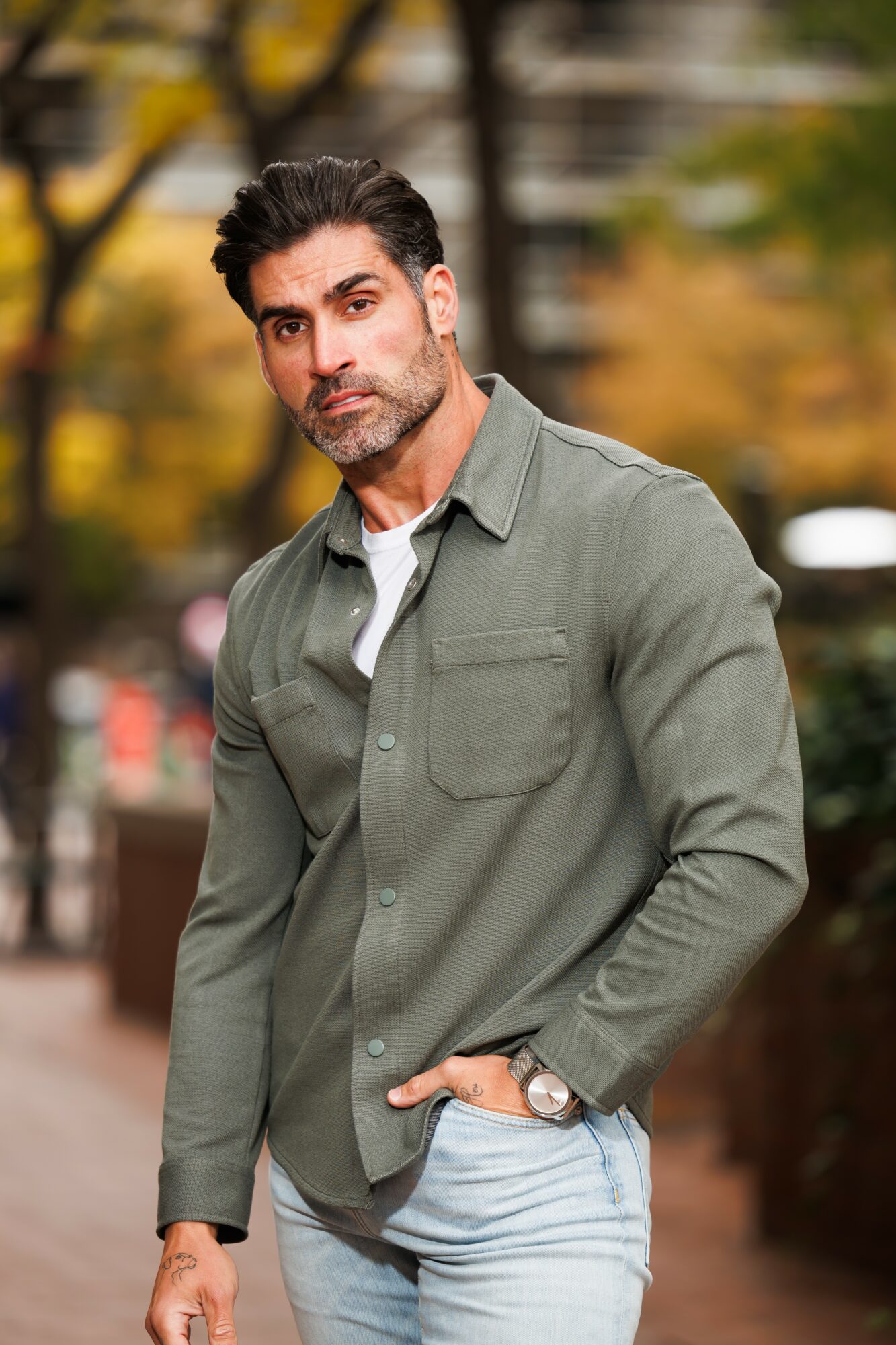 Man with dark hair and beard wearing a green jacket and white shirt outdoors, with blurred trees in background.
