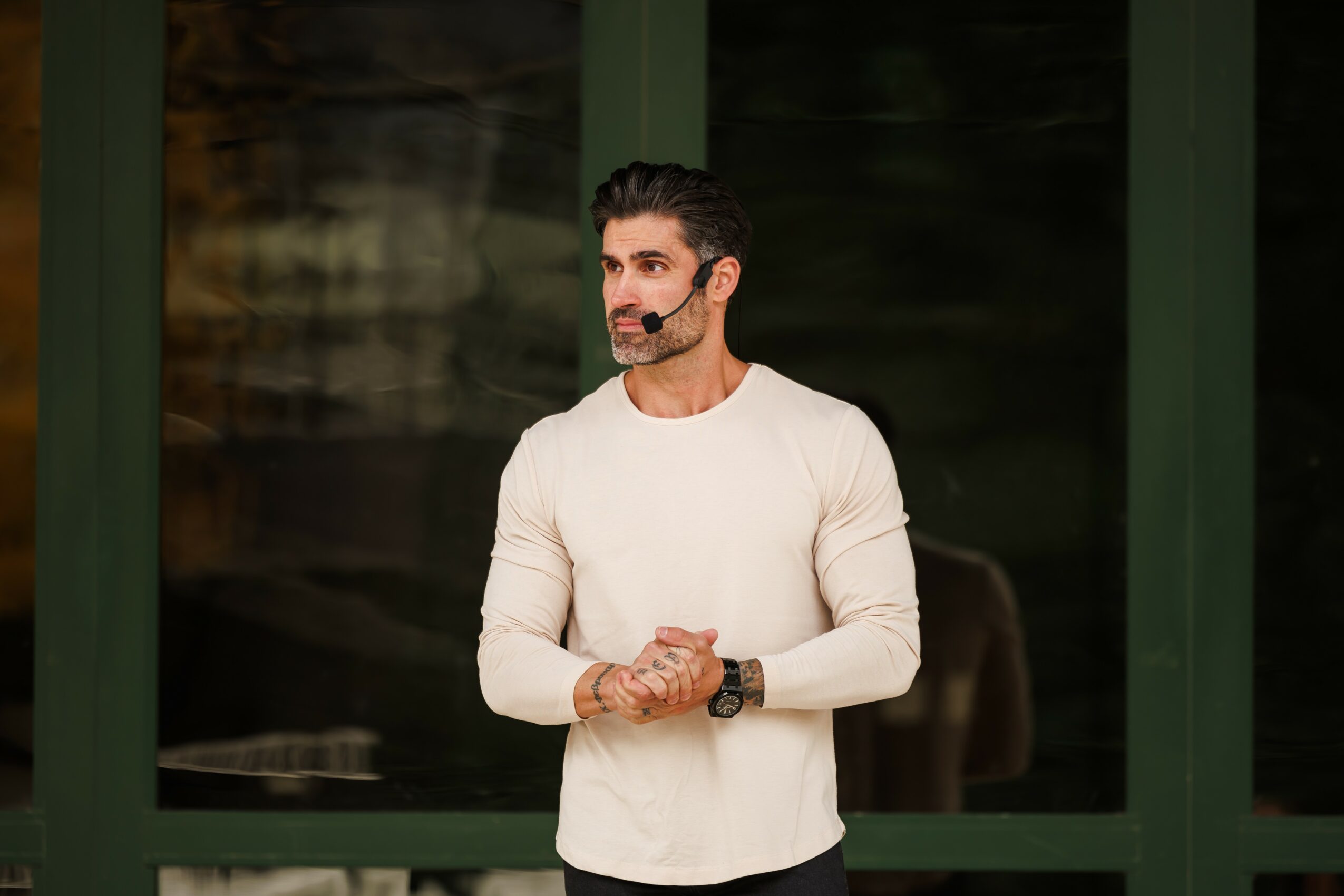 Man with dark hair and beard wearing a white long-sleeve shirt standing outdoors in front of a green-framed window.