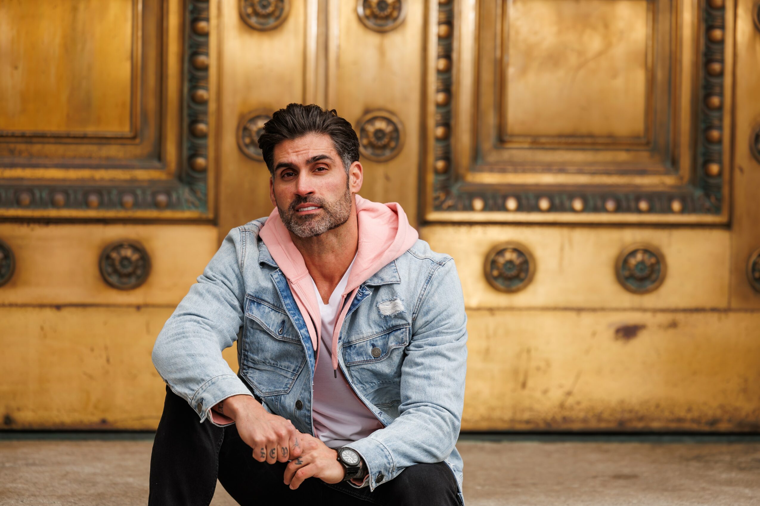 Man with dark hair and beard wearing a denim jacket and pink hoodie, sitting in front of a large wooden door with ornate details.