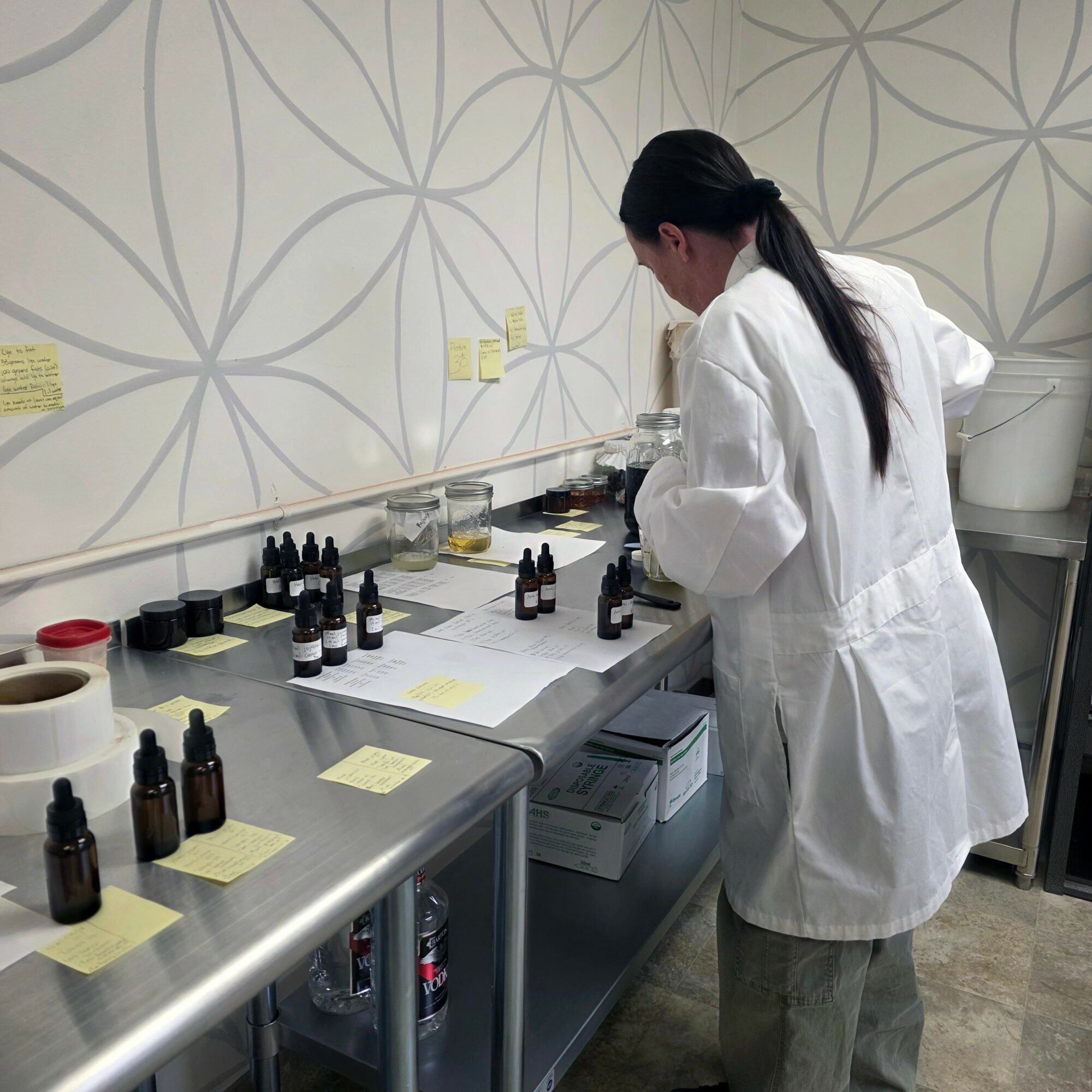 Person in a white lab coat working with small bottles and notes on a laboratory bench.