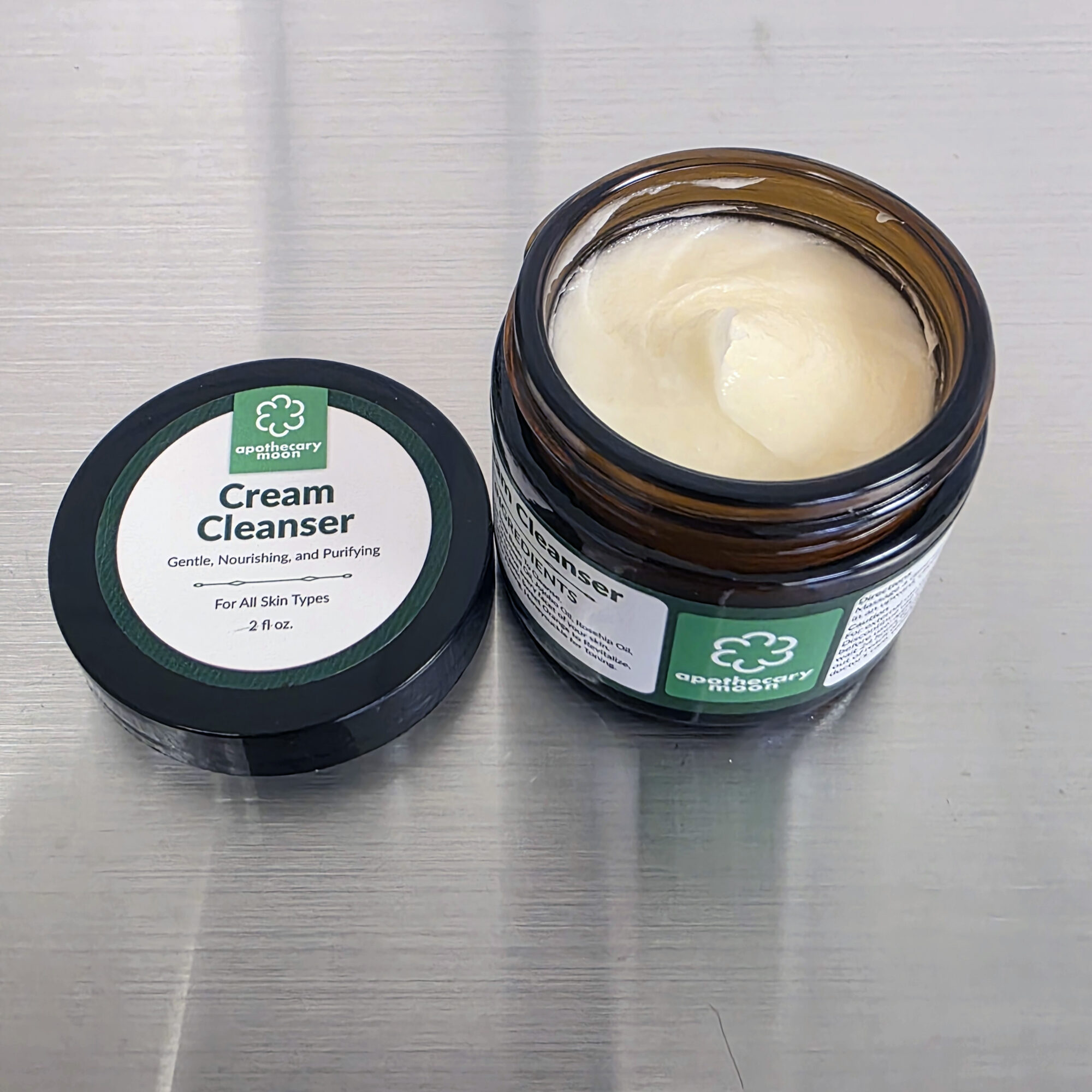 Open jar of cream cleanser with lid beside on a metallic surface.