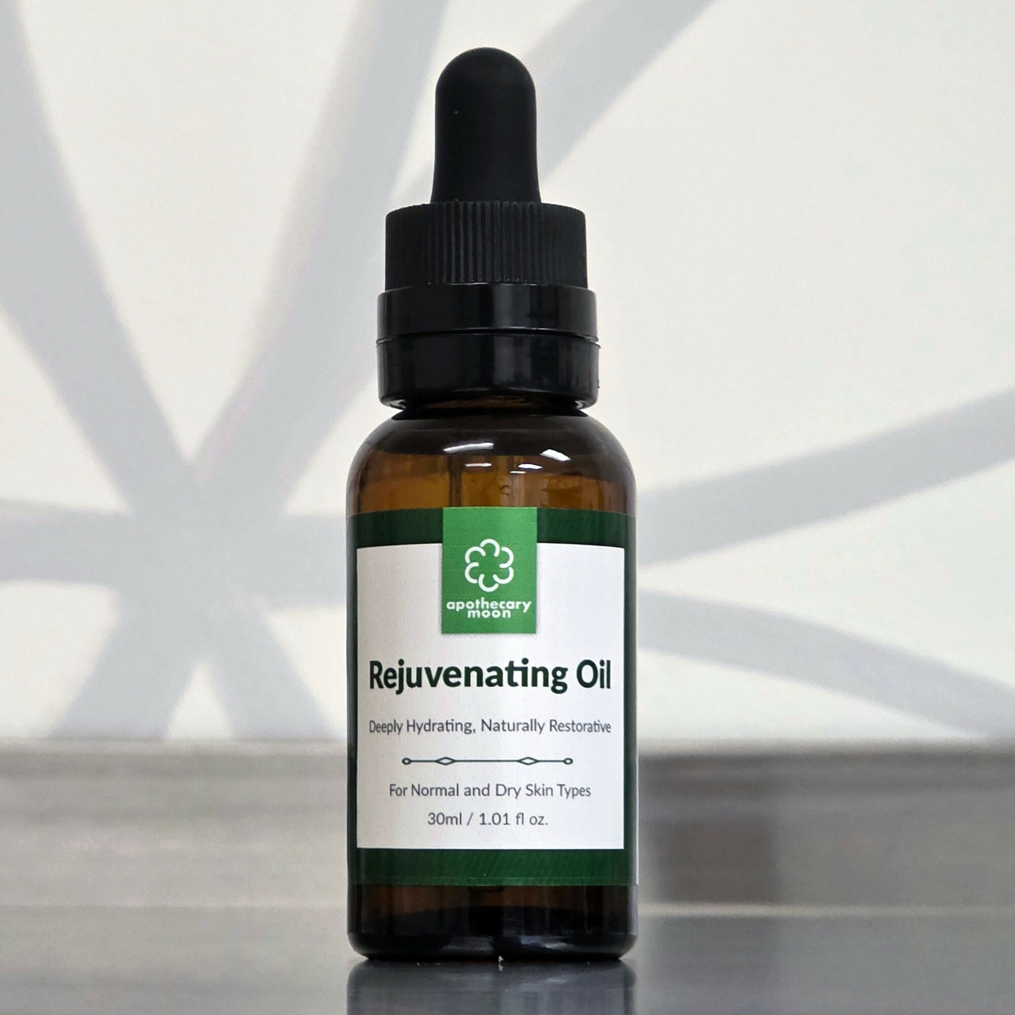 Brown glass bottle with a black dropper cap, labeled Rejuvenating Oil, on a white surface with shadows.