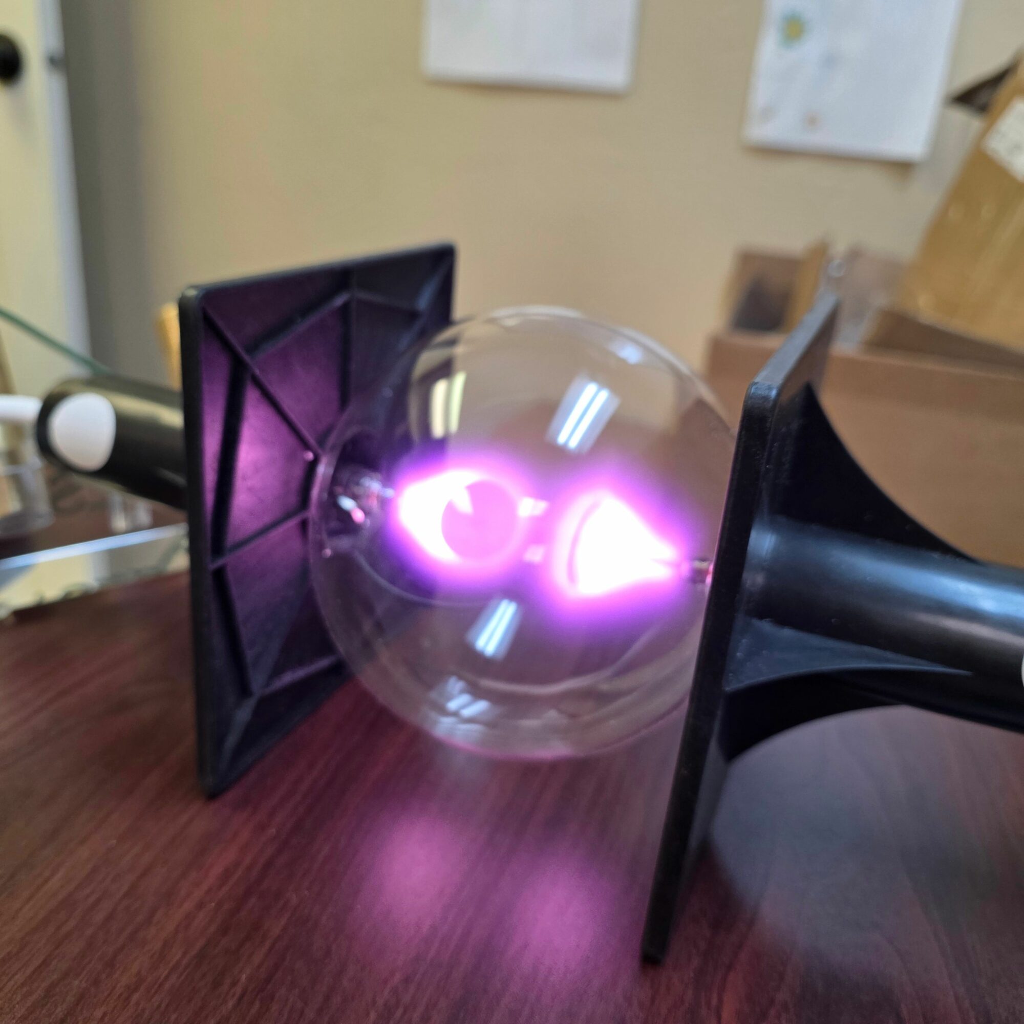 Glass bulb with purple light on a black stand on a wooden surface, background with papers on a wall.