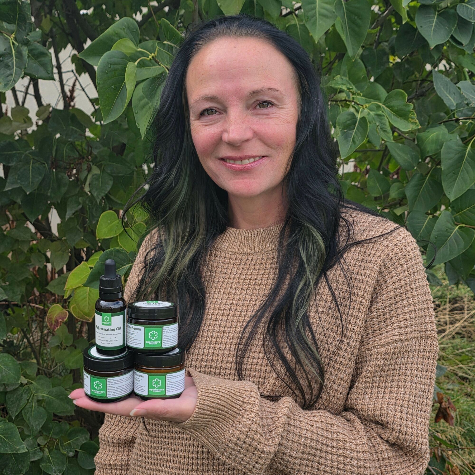 Woman with long dark hair smiling outdoors, holding three small bottles, surrounded by green foliage.