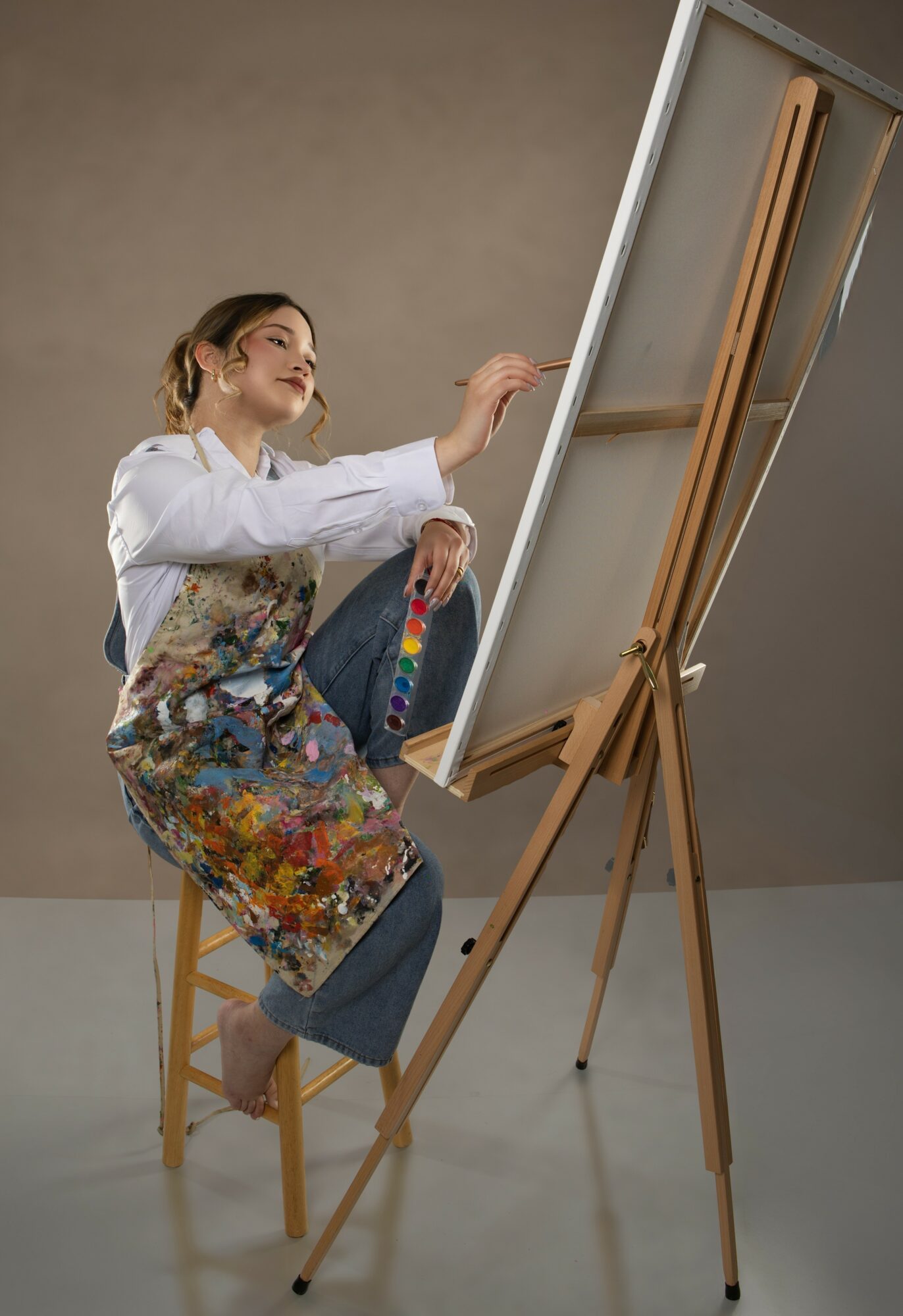 Young woman painting on a large canvas on an easel, sitting on a wooden chair, smiling, in a neutral room.