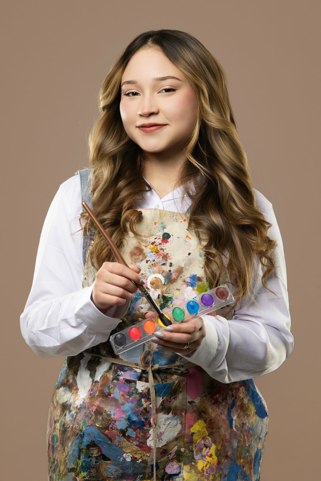 Young girl with long wavy hair holding a paint palette and brush, wearing a white shirt and apron, smiling.