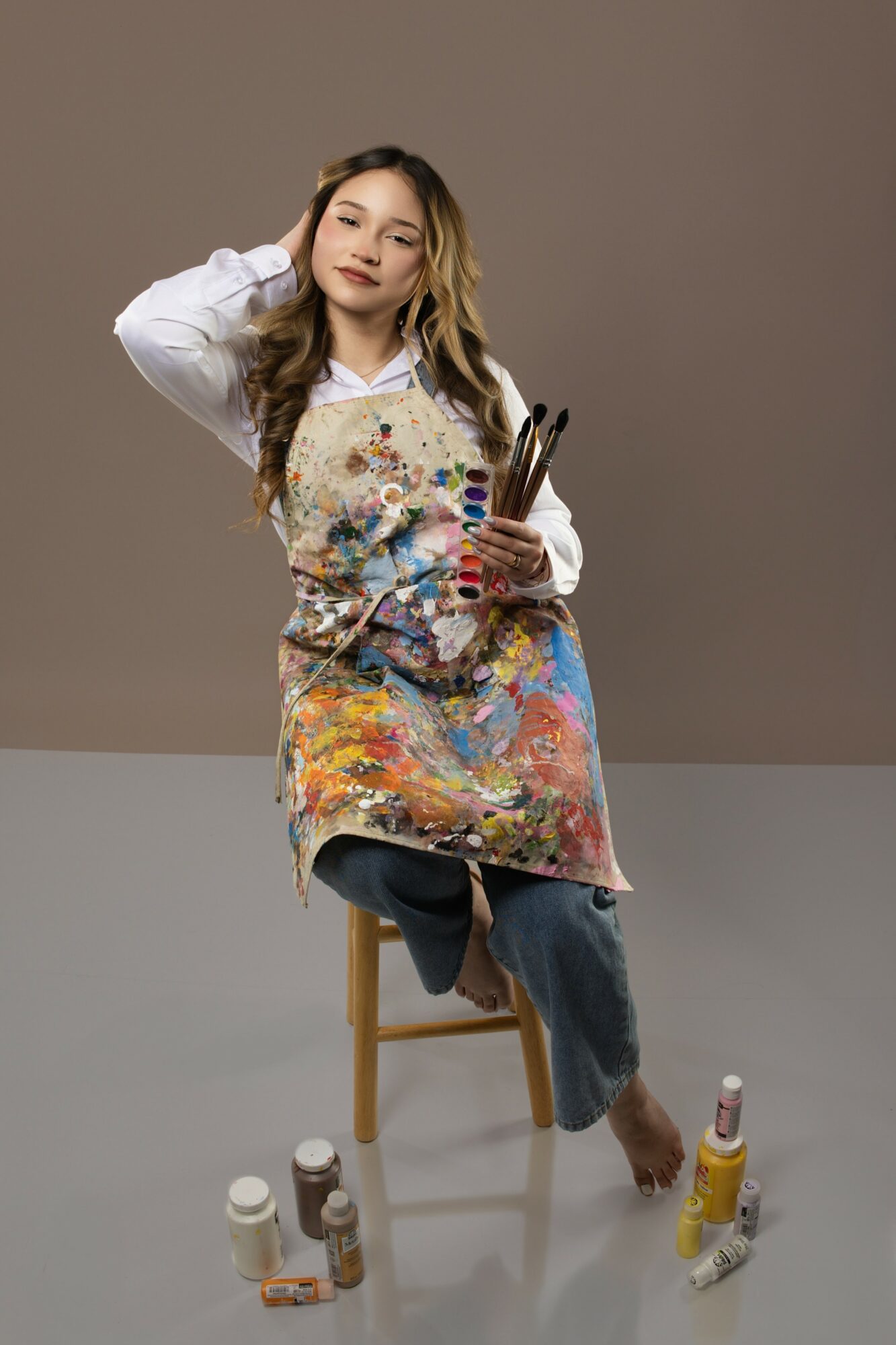 Young woman sitting on a stool, holding paintbrushes and palette, wearing a colorful apron and casual clothing, against a plain background.