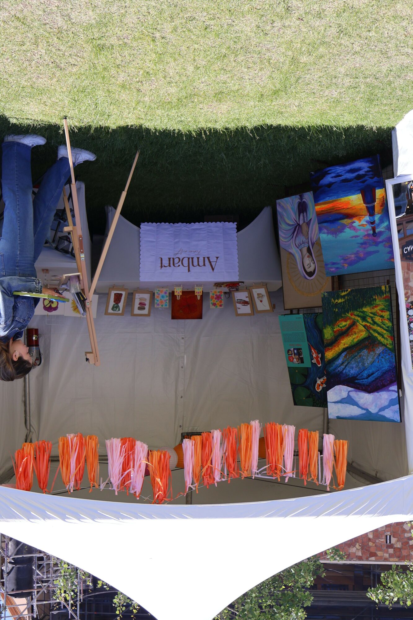 Outdoor art display with colorful paintings and fabric hanging, table with art supplies, and a person working on art.