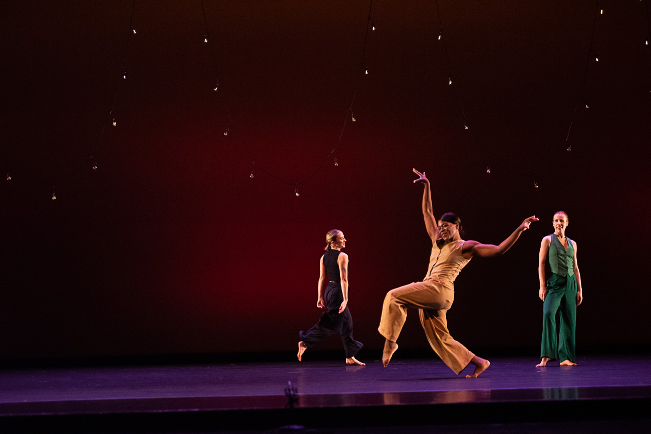 Three dancers perform on stage with a dark background and star-like lights, one in a dynamic pose, two standing still.