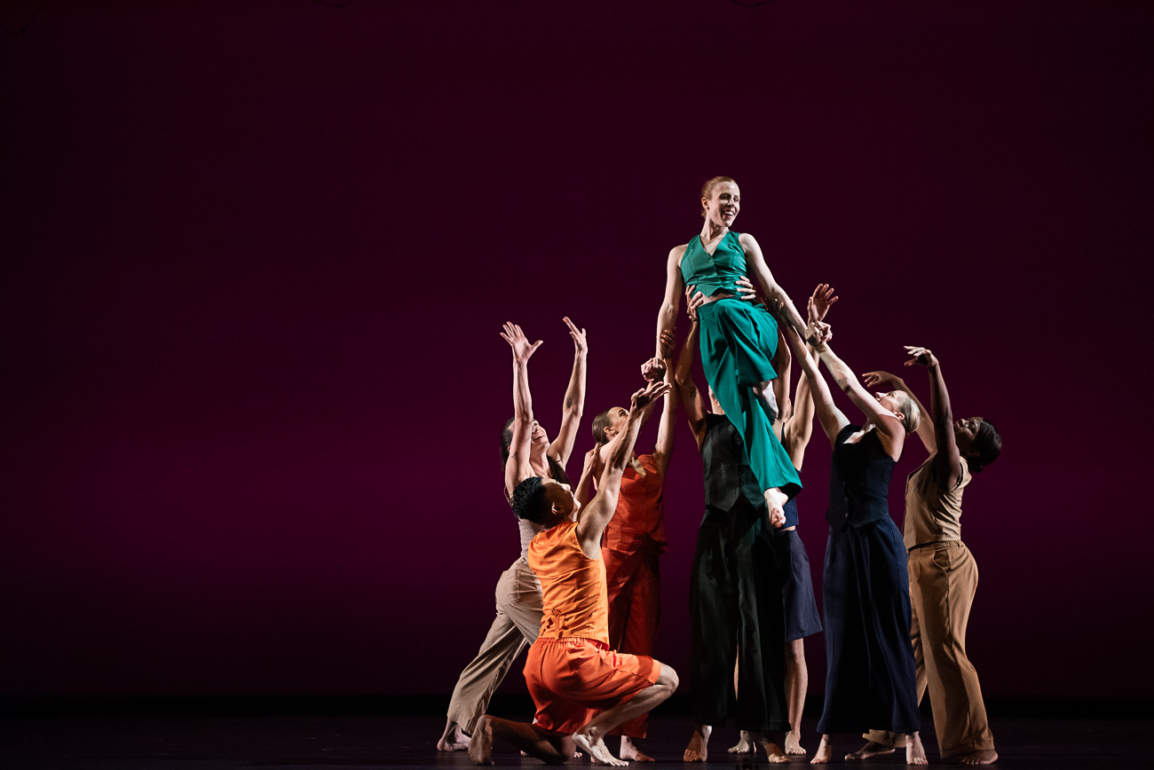 A performer in a teal dress is lifted by dancers on stage with a dark background.