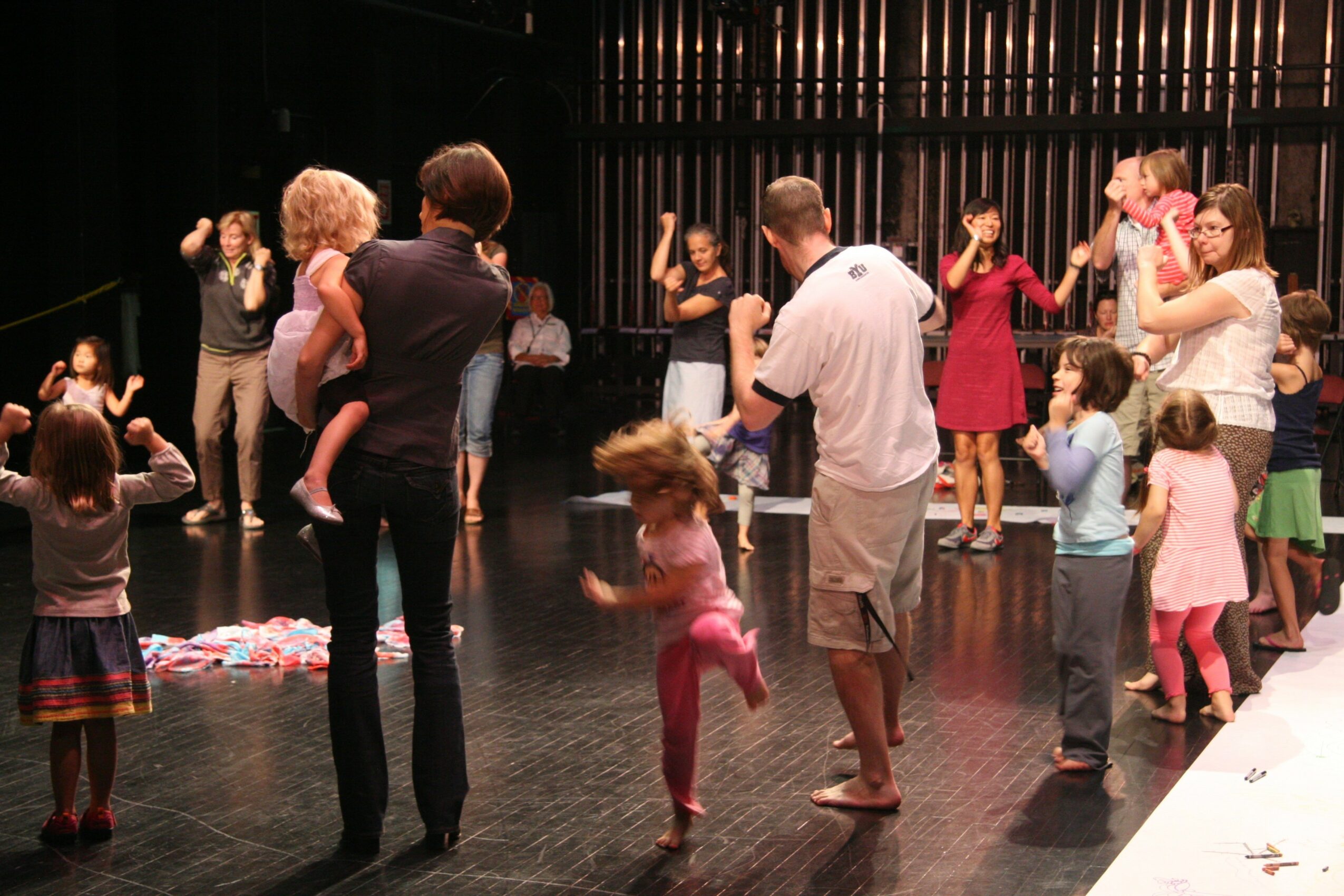 Group of children and adults dancing and playing on a stage with a dark background, some holding hands and jumping.