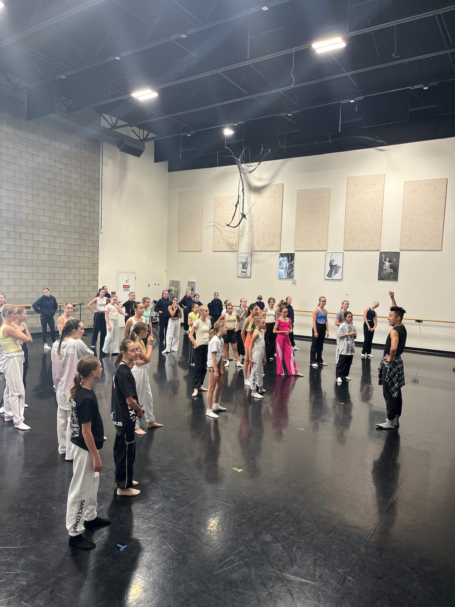 Group of children and adults standing in a spacious dance studio with high ceiling and black floor.